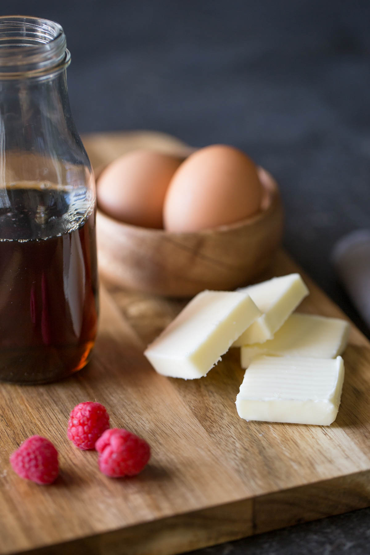 A cutting board with four pads of butter, a small wood bowl of brown eggs, a glass jar of maple syrup and a few fresh raspberries. 