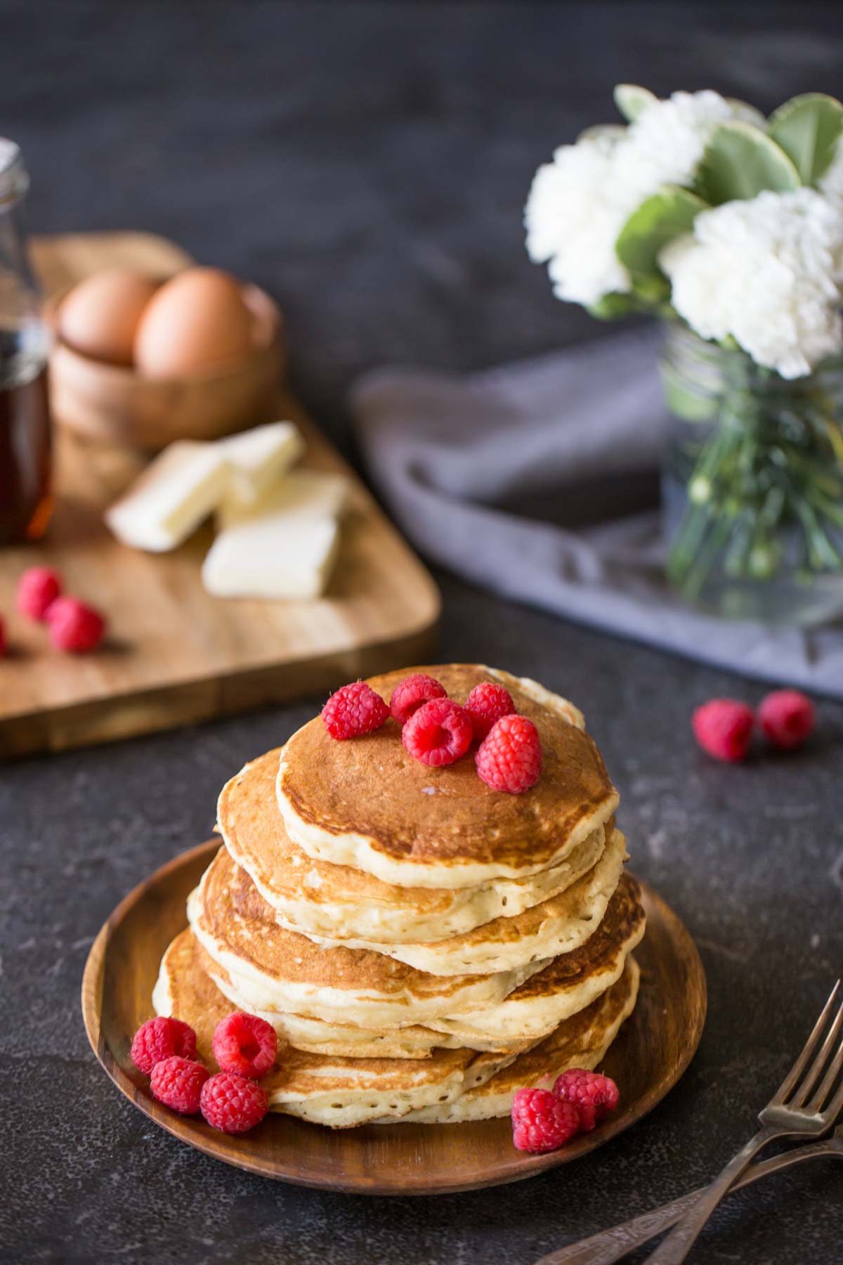 A stack of the Best Ever Buttermilk Pancakes on a plate, topped with fresh raspberries, with a cutting board with four pads of butter, a small wood bowl of brown eggs, a glass jar of maple syrup and a few fresh raspberries in the background, along with a flower arrangement. 