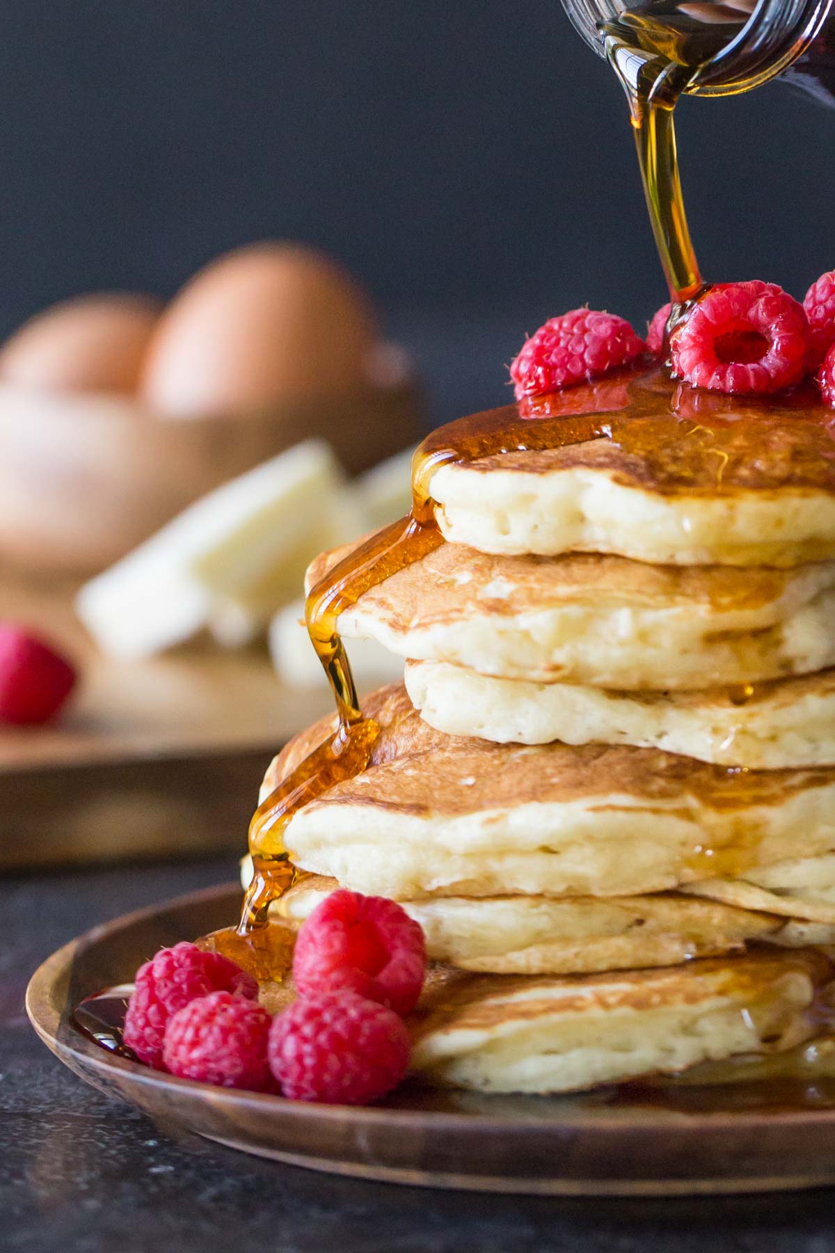A stack of the Best Ever Buttermilk Pancakes on a plate, topped with fresh raspberries, with maple syrup being poured over them. 