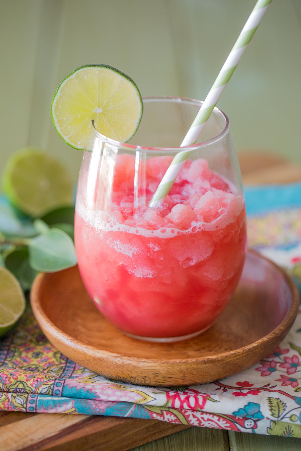 Coconut Lime Watermelon Slushie in a glass tumbler with a straw and a slice of lime on the rim, sitting on a small wood plate. 