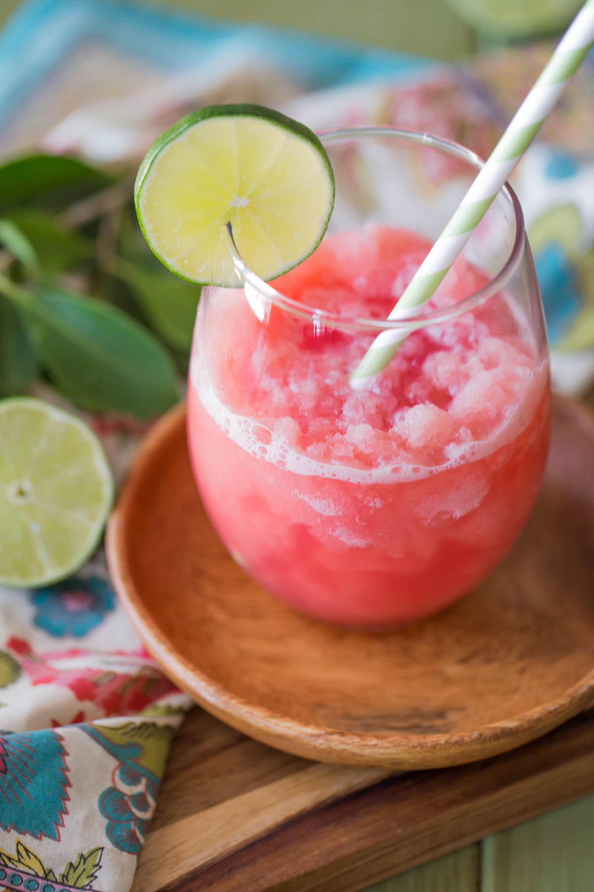 Coconut Lime Watermelon Slushie in a glass tumbler with a straw and a slice of lime on the rim, sitting on a small wood plate. 
