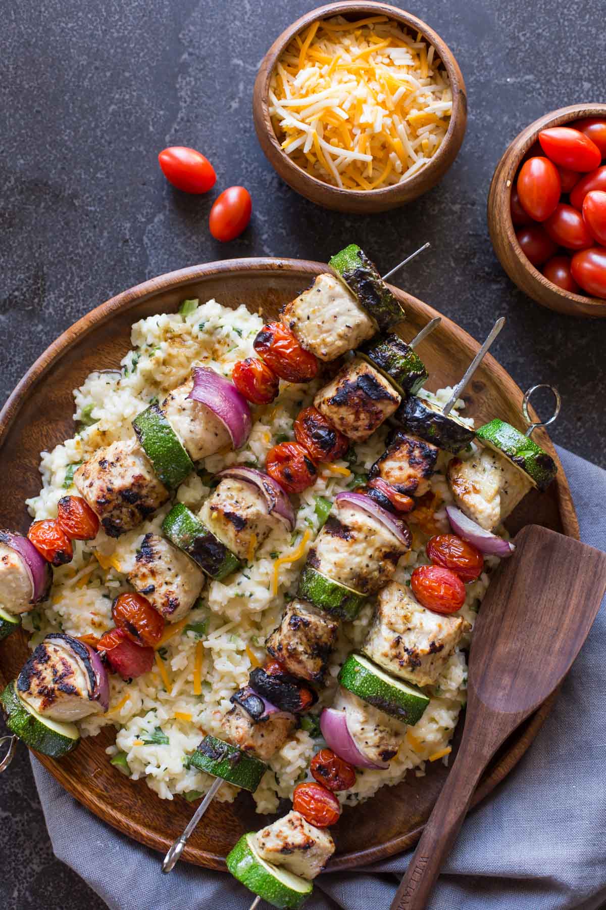Pork Tenderloin Kabobs served over Cheesy Broccoli Rice on a large wood plate, with a small wood bowl of shredded cheese and a small wood bowl of grape tomatoes next to it. 