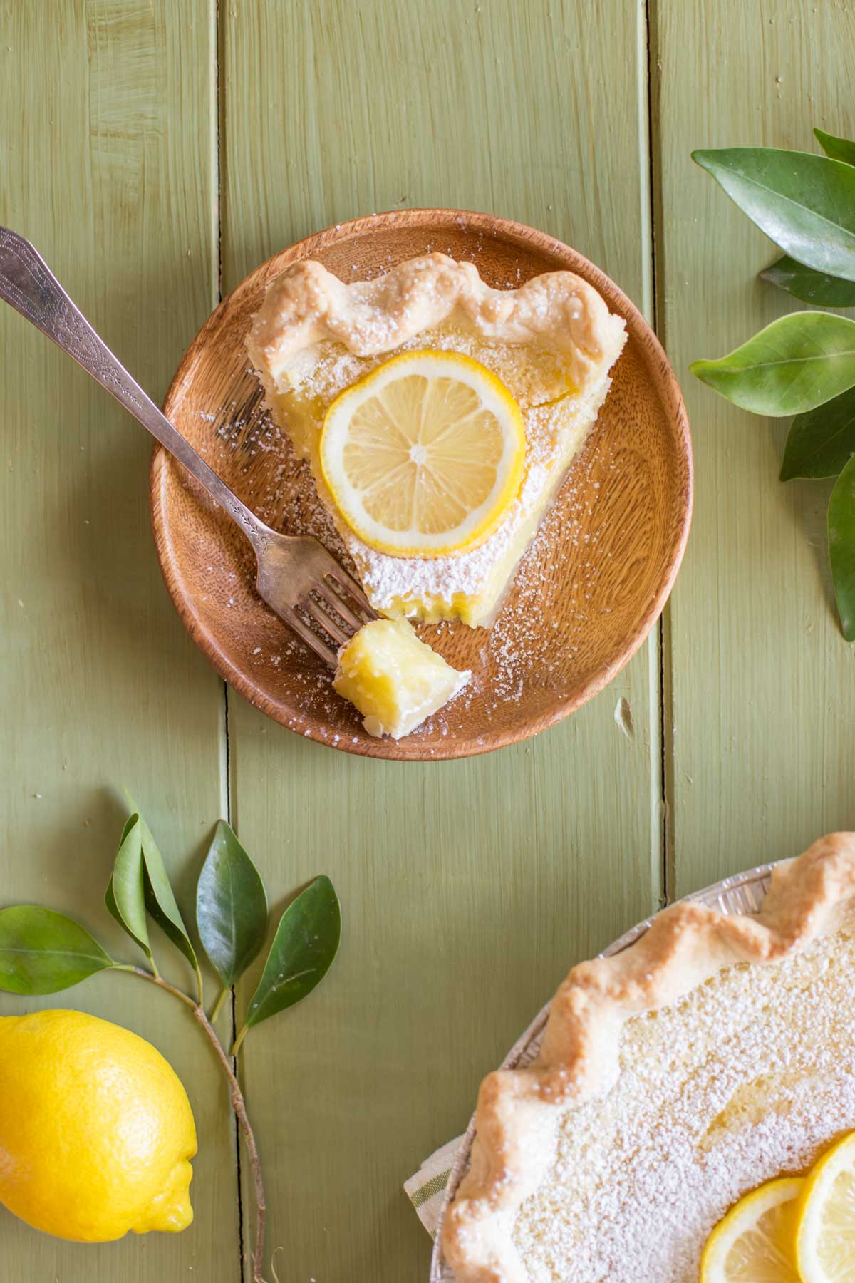 A slice of Whole Lemon Pie dusted with powdered sugar and topped with a lemon slice, on a plate with a fork with a piece of pie on it, siting next to the Whole Lemon Pie pan. 