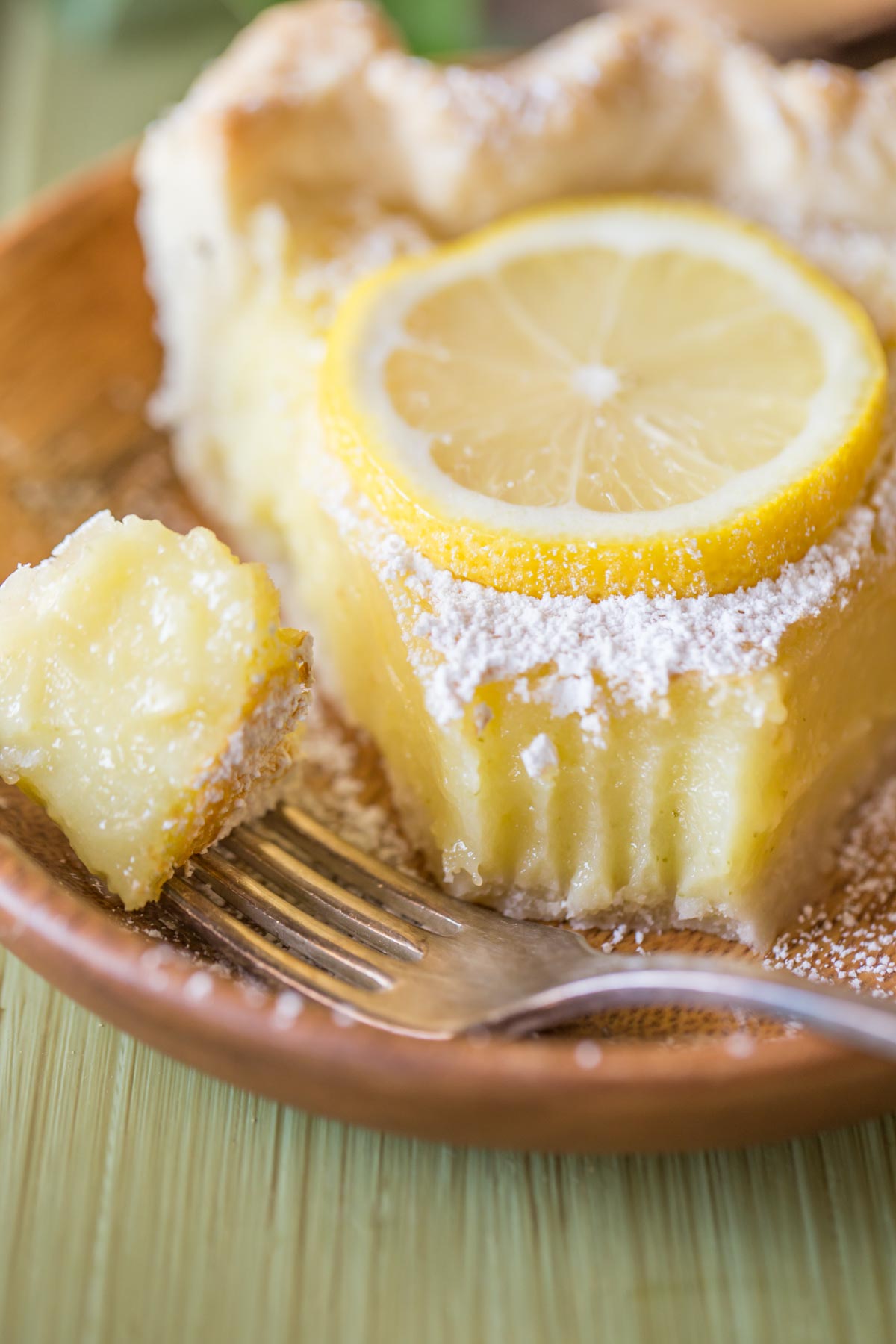 A slice of Whole Lemon Pie dusted with powdered sugar and topped with a lemon slice, on a plate with a fork with a piece of pie on it. 