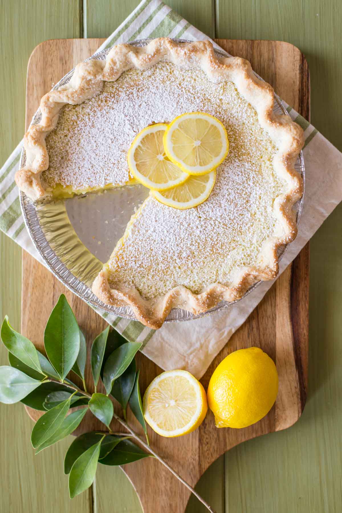 A Whole Lemon Pie dusted with powdered sugar and garnished with lemon slices, with one slice of pie missing, sitting on a cutting board with some lemons and tree leaves. 
