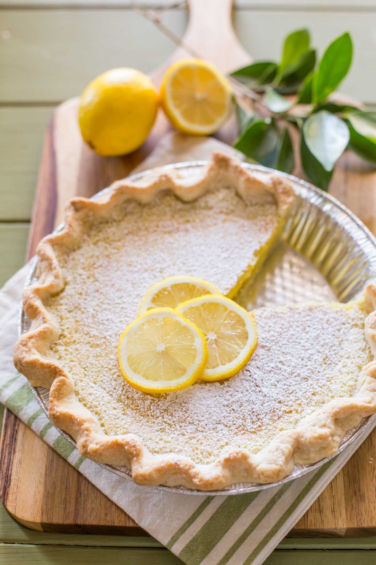 A Whole Lemon Pie dusted with powdered sugar and garnished with lemon slices, with one slice of pie missing, sitting on a cutting board with some lemons and tree leaves. 