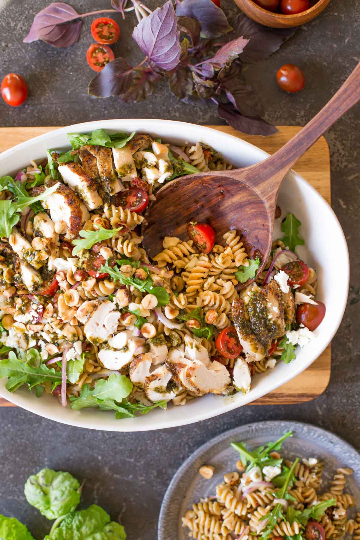 Whole Wheat Pasta Salad With Honey Basil Balsamic Vinaigrette in a large serving bowl with a wooden spoon in it, and a serving of the pasta salad on a plate next to it. 