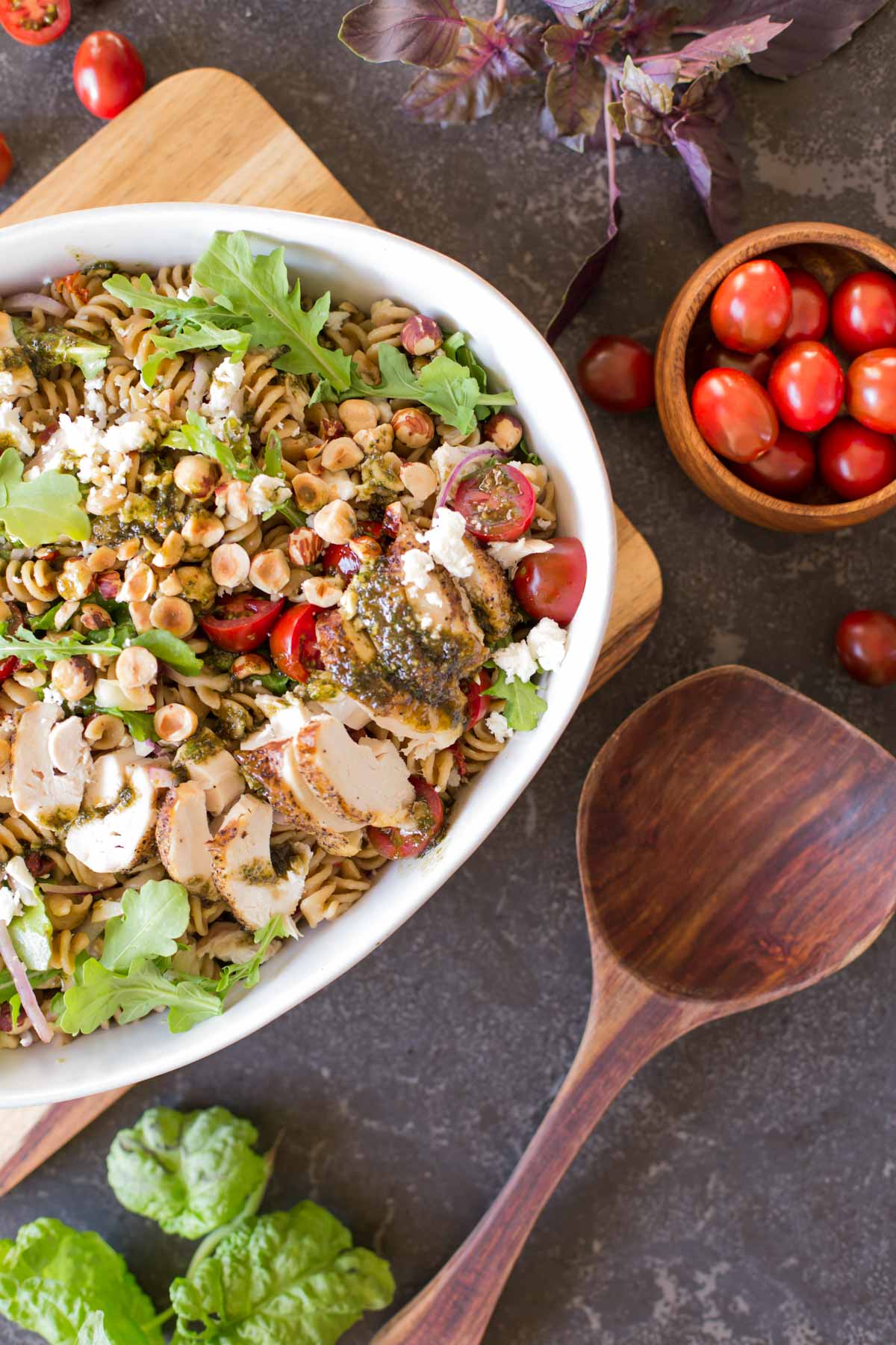 Whole Wheat Pasta Salad With Honey Basil Balsamic Vinaigrette in a large serving bowl with a wooden spoon next to it, as well as a small wood bowl of grape tomatoes. 
