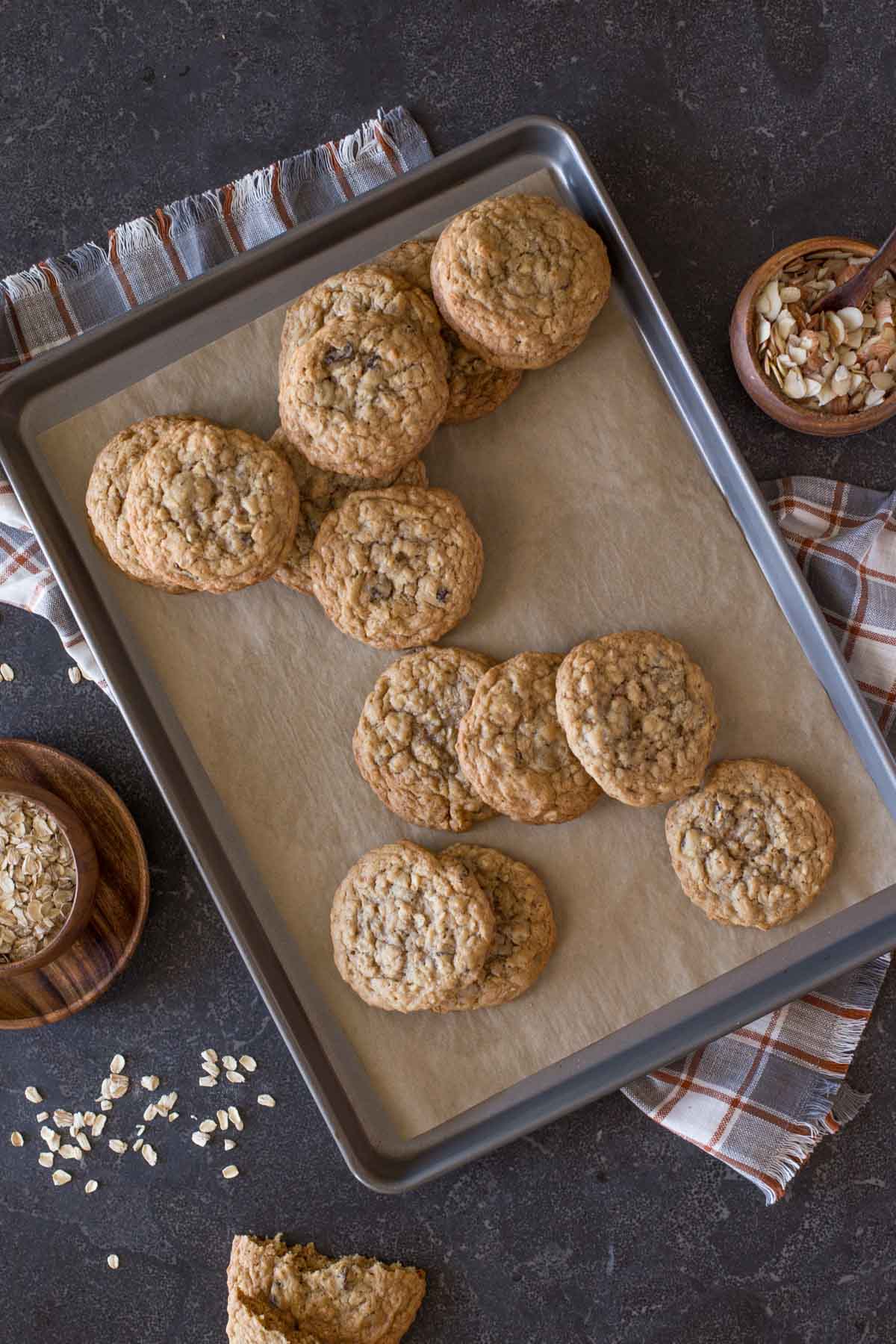Bakery Style Oatmeal Raisin Cookies on a parchment paper lined baking sheet, with a small wood bowl of oats on one side of the baking sheet, and a small wood bowl of slice almonds on the other side. 