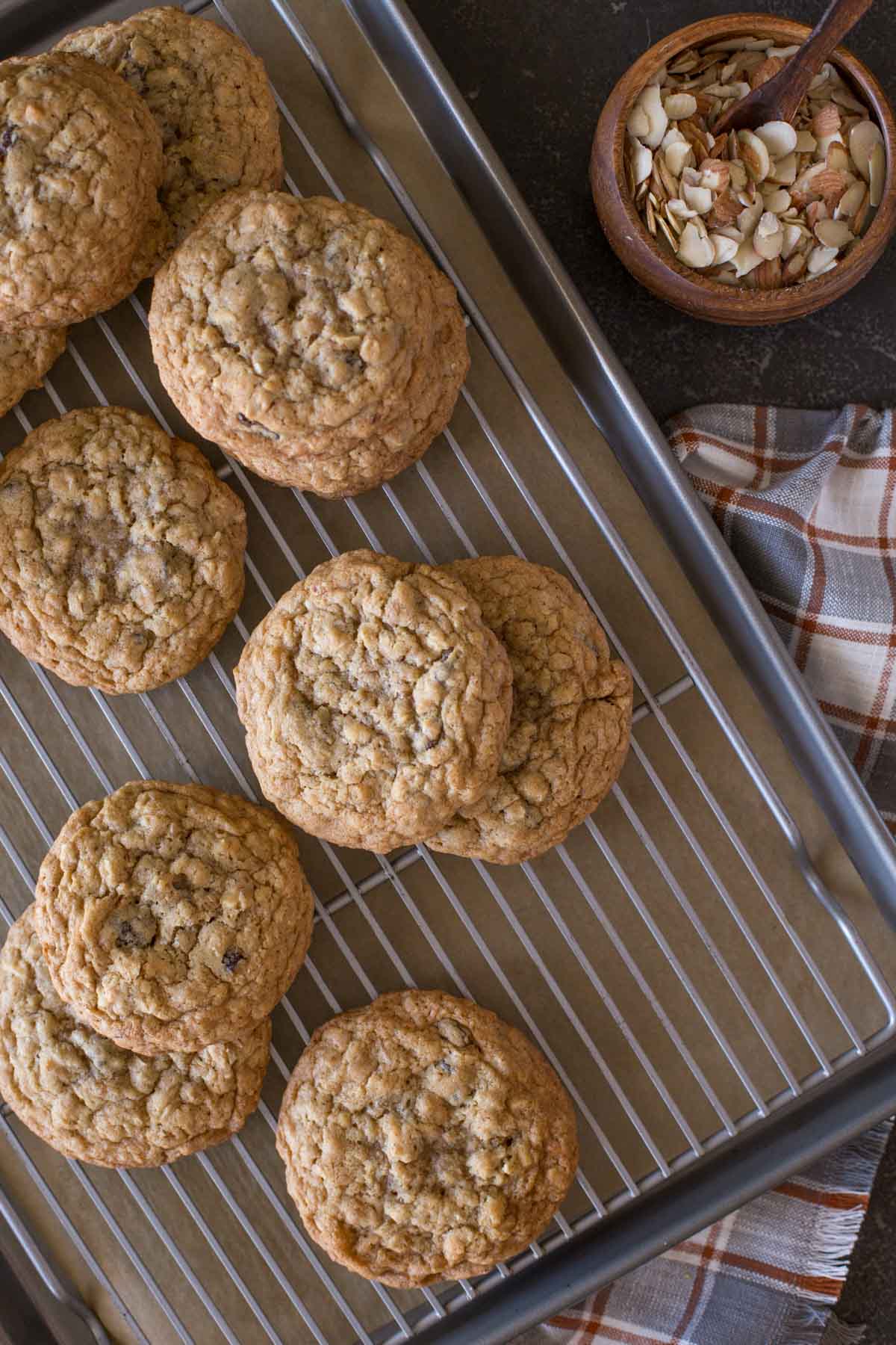 Bakery Style Oatmeal Raisin Cookies on a cooling rack that is on top of a parchment paper lined baking sheet, with a small wood bowl of slice almonds next to it. 