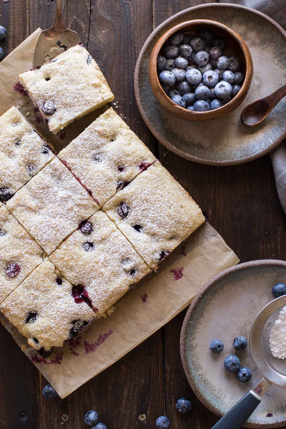 Buttermilk Blueberry Snack Cake sliced on a piece of parchment paper, with a small wood bowl of blueberries on a plate next to it, as well as a plate with a sifter full of powdered sugar.