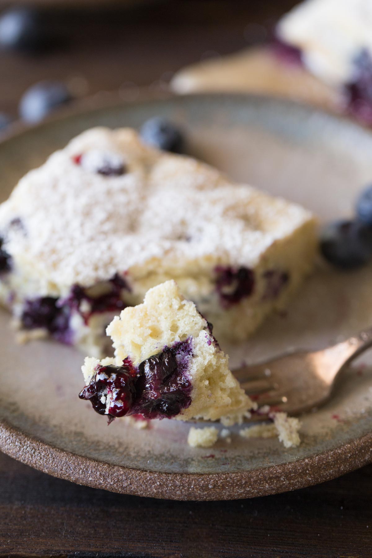 A piece of Buttermilk Blueberry Snack Cake on a plate, with a fork with a bite of cake on it. 