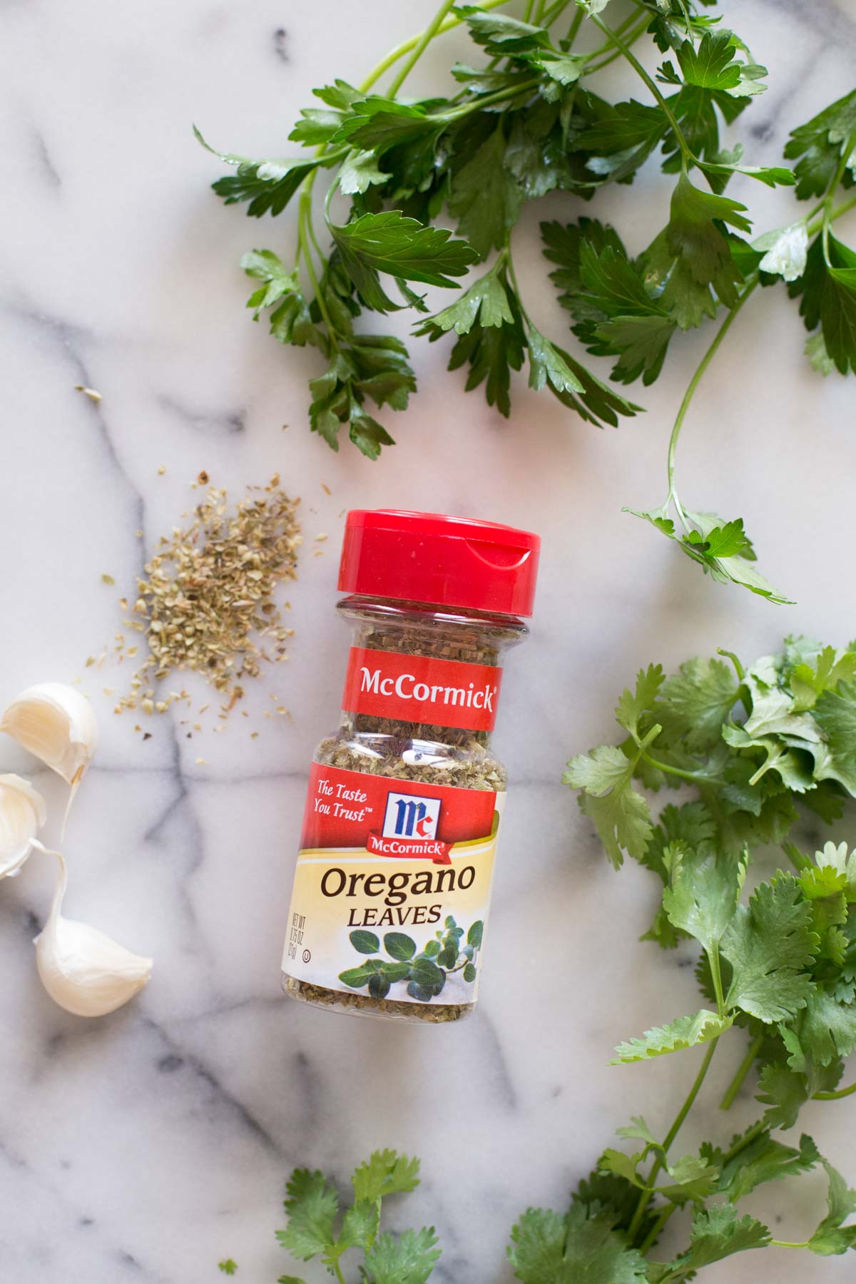 A spice container of McCormick Oregano Leaves, fresh parsley, cilantro, and garlic for the chimichurri.