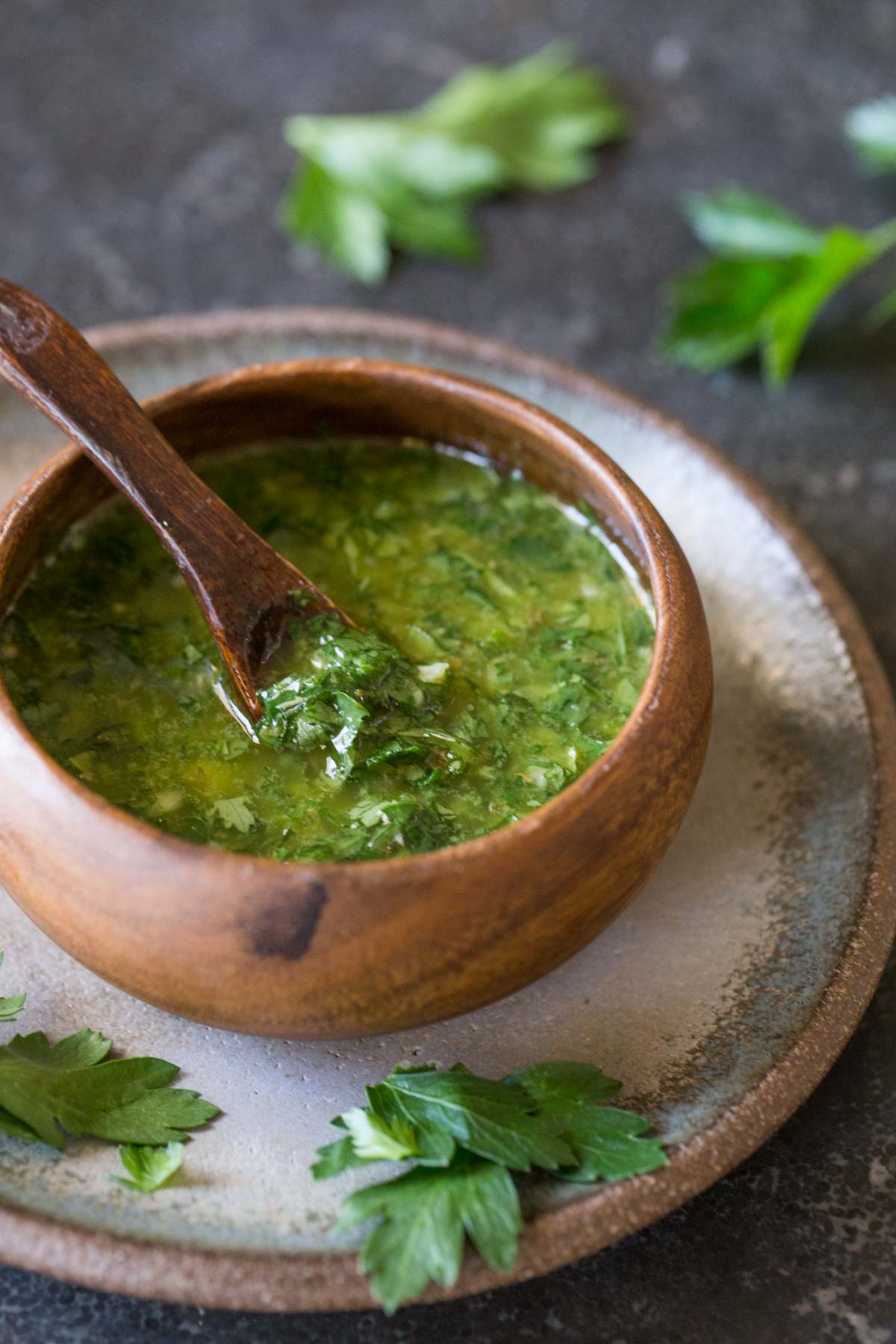 Chimichurri in a small wood bowl with a wooden spoon it it, sitting on a plate. 