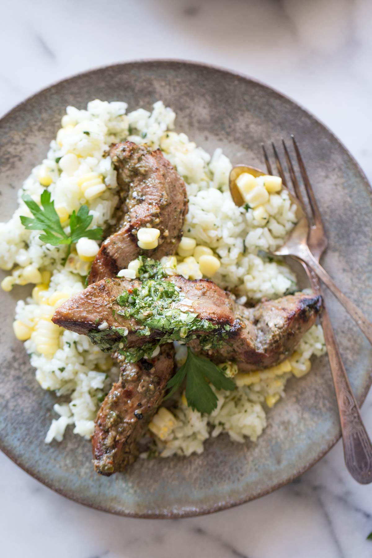 Brazilian Grilled Flank Steak resting on a bed of Chimichurri Rice, on a plate with a fork and a spoon.