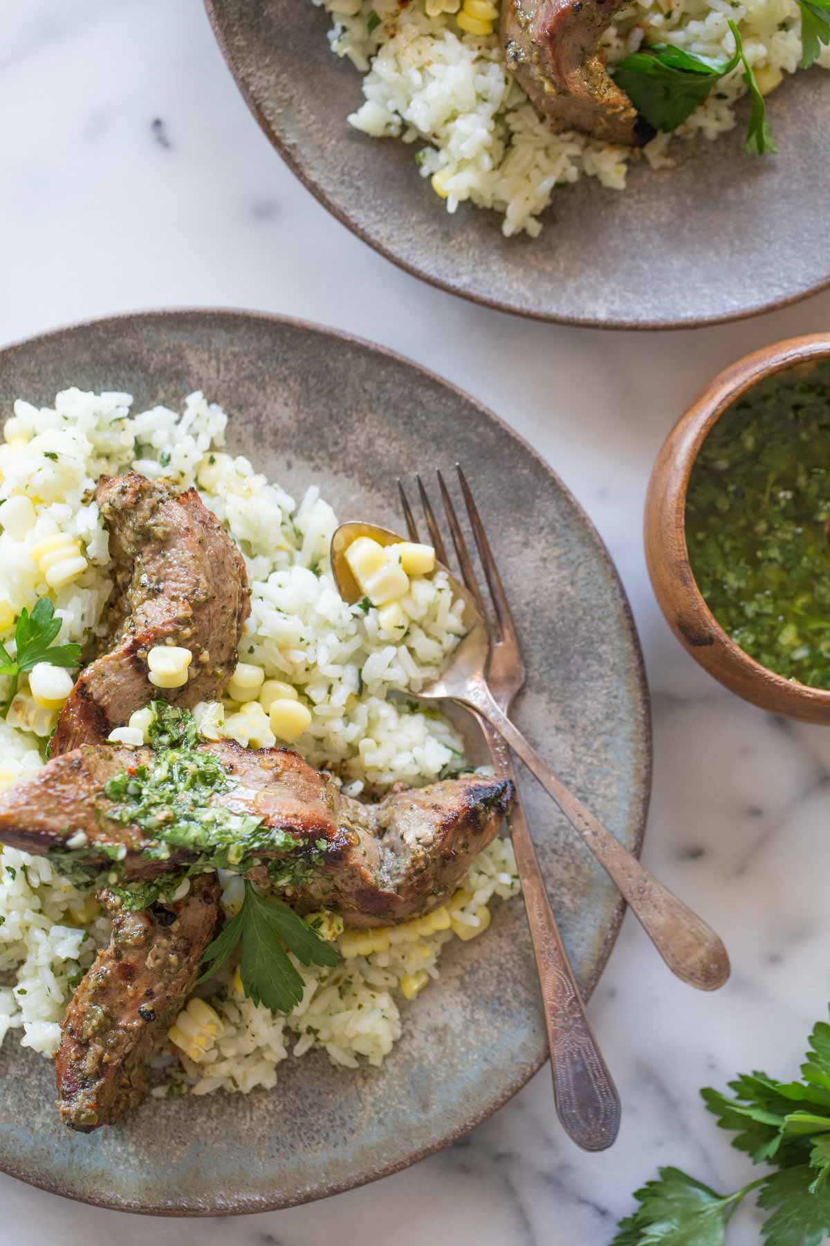 Brazilian Grilled Flank Steak resting on a bed of Chimichurri Rice, on a plate with a fork and a spoon, with another plate of Brazilian Grilled Flank Steak resting on a bed of Chimichurri Rice next to it, as well as a small bowl of chimichurri. 