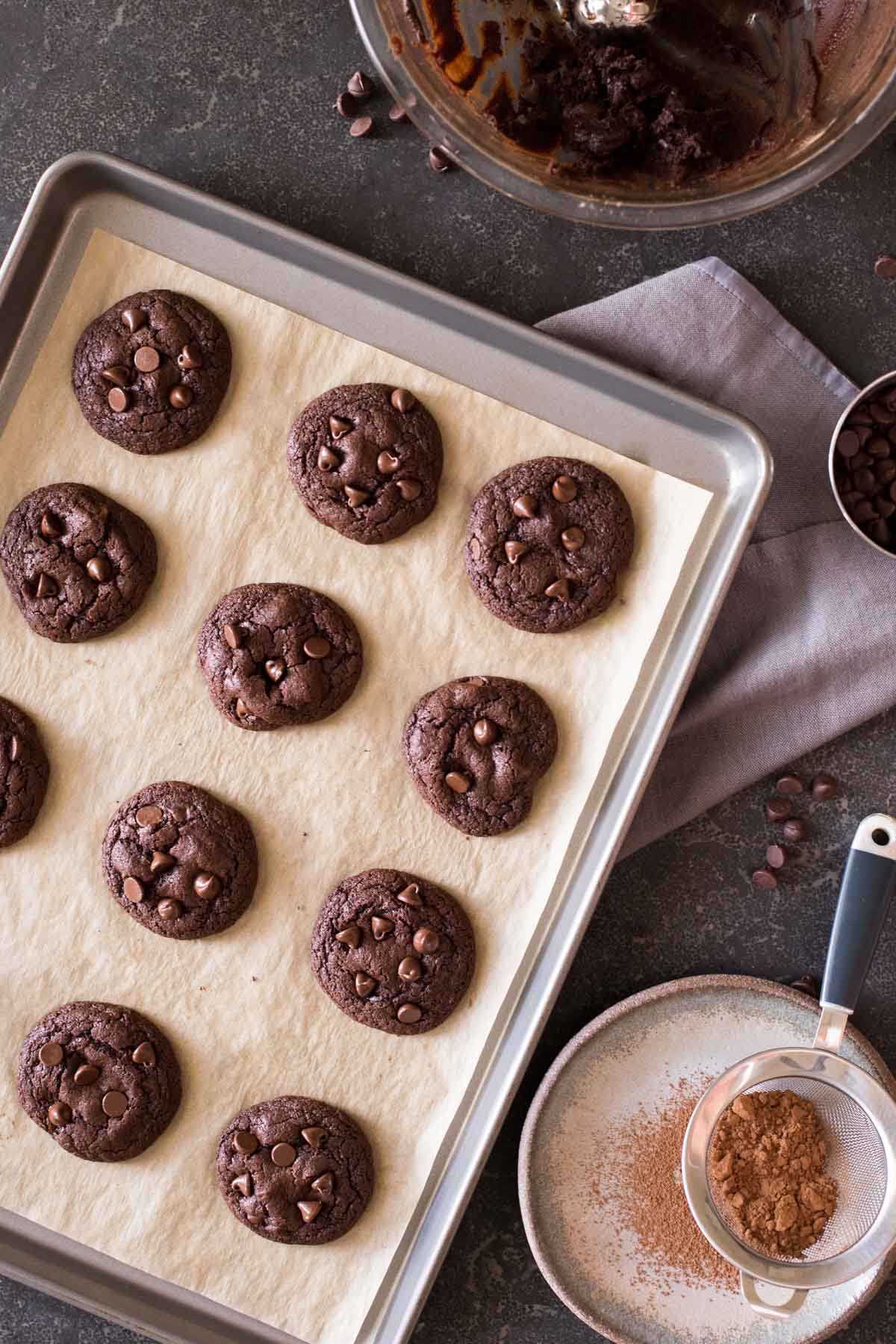 One Bowl Triple Chocolate Brownie Cookies on a parchment paper lined baking sheet, sitting next to the bowl of cookie dough and a plate with a little sifter with cocoa powder in it. 