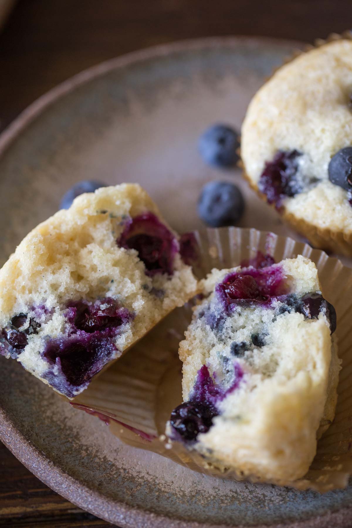 A Vanilla Blueberry Buttermilk Muffin split in half, sitting on a plate with another whole muffin and some blueberries. 