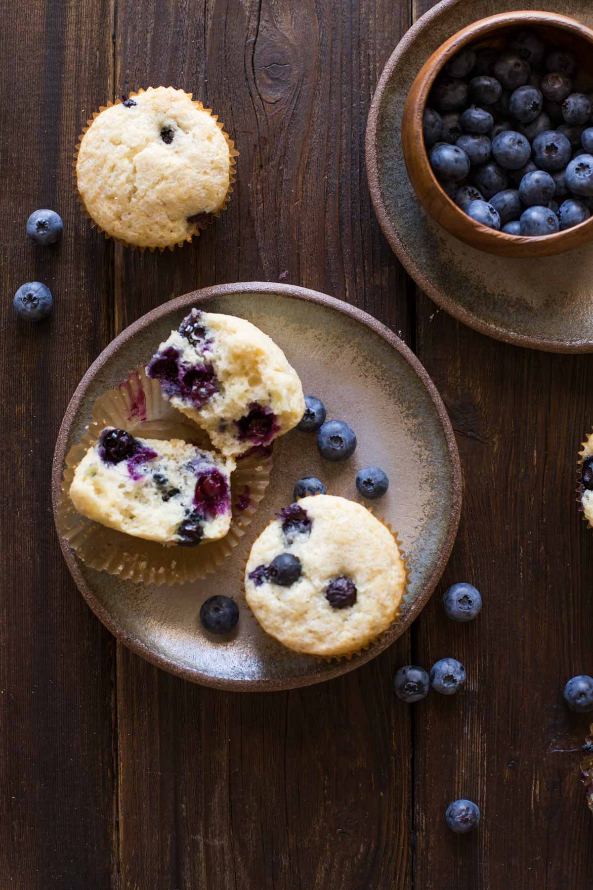 A Vanilla Blueberry Buttermilk Muffin split in half, sitting on a plate with a whole muffin and some blueberries, with a muffin and blueberries near the plate, and a wood bowl of blueberries sitting on another plate. 