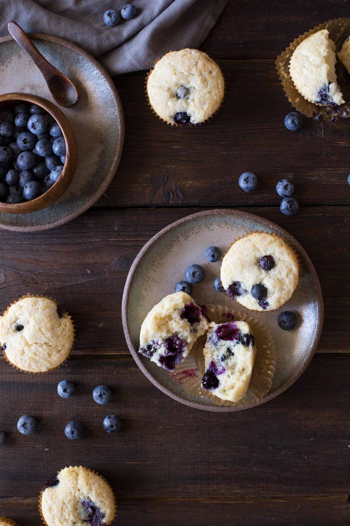 A Vanilla Blueberry Buttermilk Muffin split in half, sitting on a plate with a whole muffin and some blueberries, with more muffins and blueberries near the plate, and a wood bowl of blueberries sitting on another plate. 