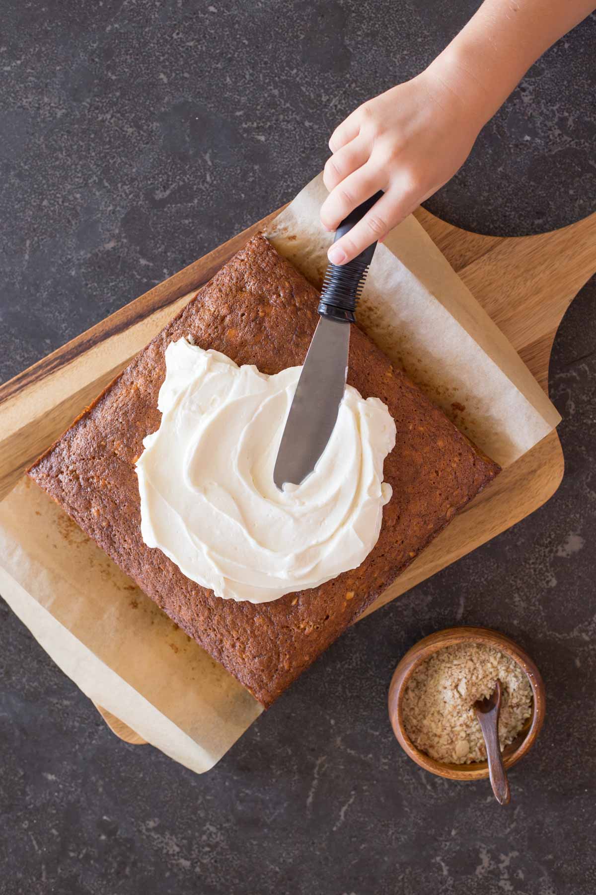 Banana Cake on parchment paper on top of a cutting board, with the Fluffy Cream Cheese Frosting being spread on top of the cake, and a small wood bowl of crushed almonds sitting next to the cutting board. 