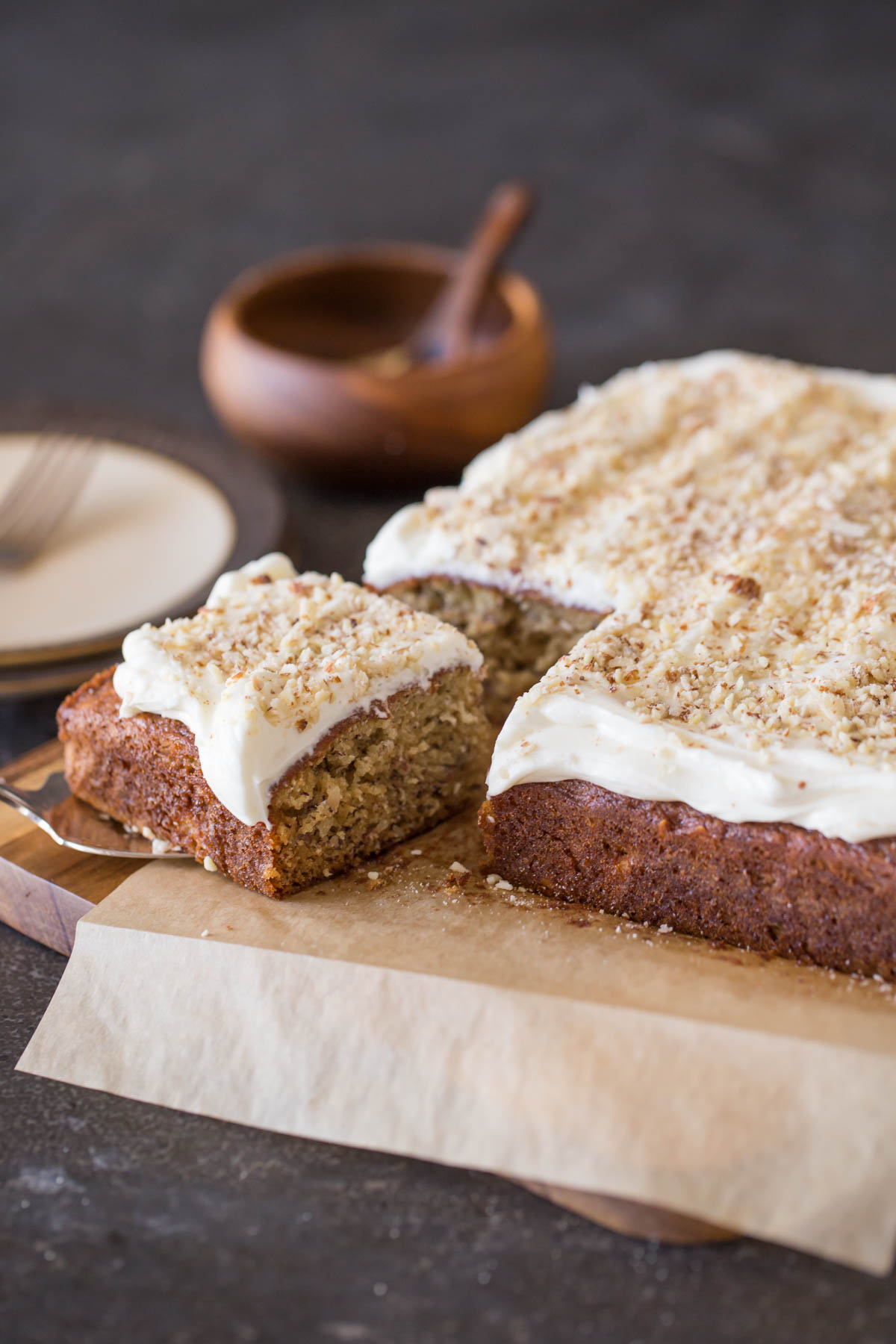 Banana Cake with Fluffy Cream Cheese Frosting on parchment paper on top of a cutting board, with the corner piece on top of a serving spatula. 