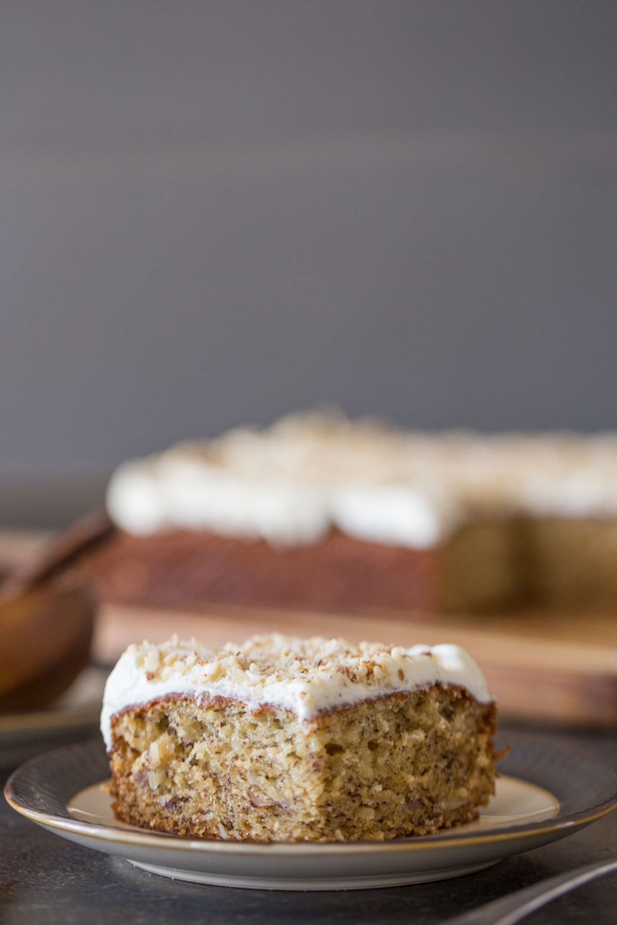 A piece of Banana Cake with Fluffy Cream Cheese Frosting on a plate, with the rest of the cake in the background. 