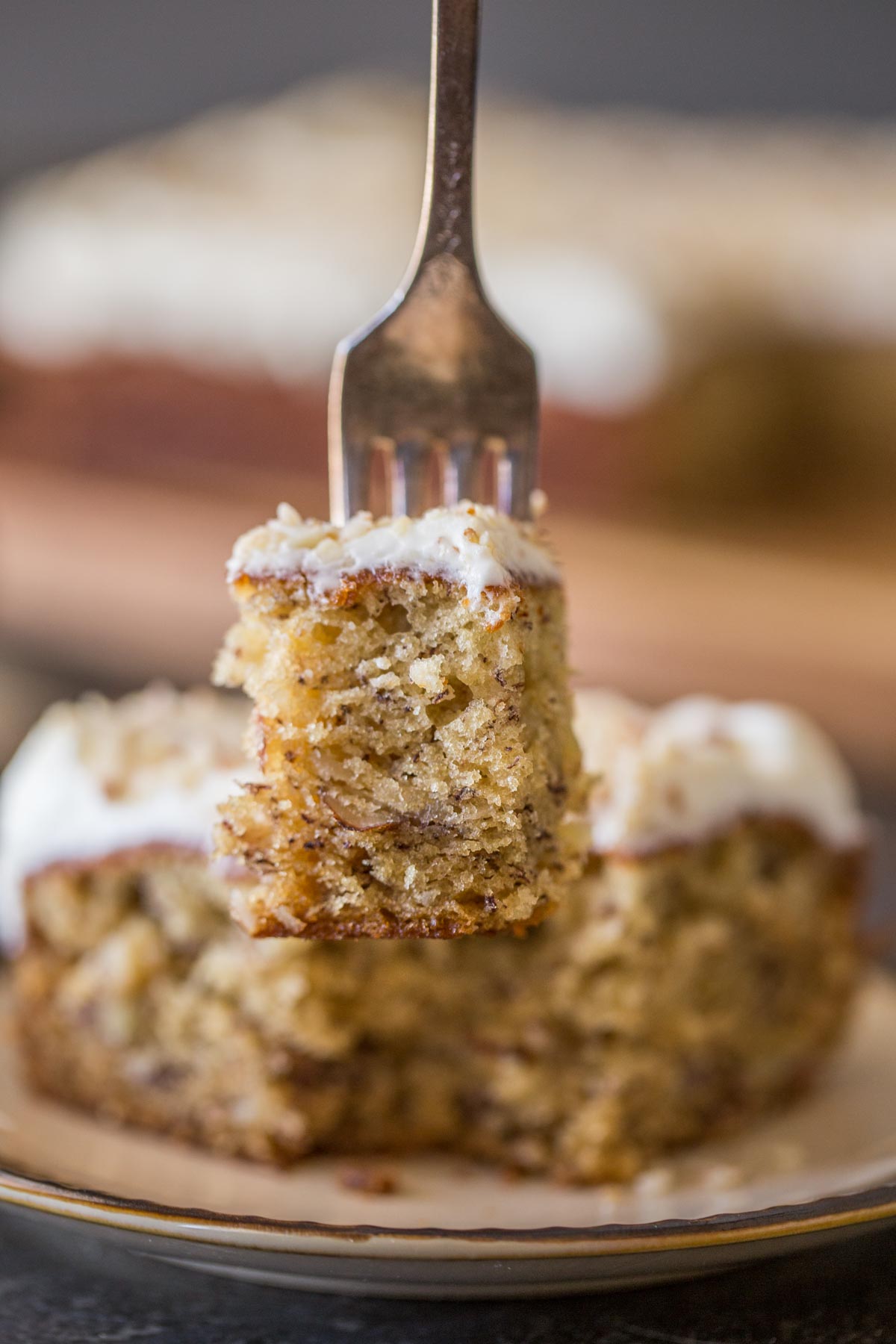 A fork with a bite of the Banana Cake with Fluffy Cream Cheese Frosting on it, with the rest of the piece of cake on a plate in the background.