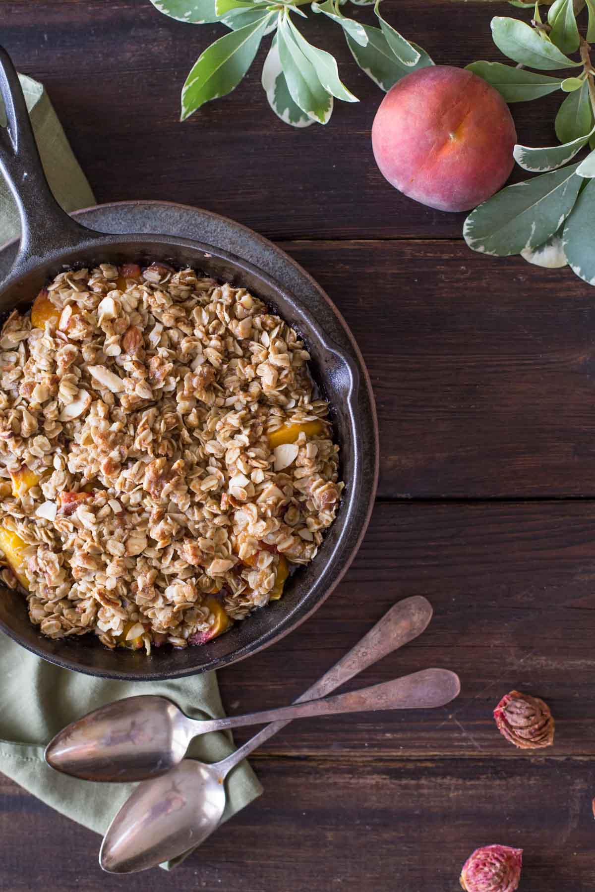 Easy Skillet Peach Crisp, with two spoons next to the skillet, as well as a whole peach and a couple peach pits. 