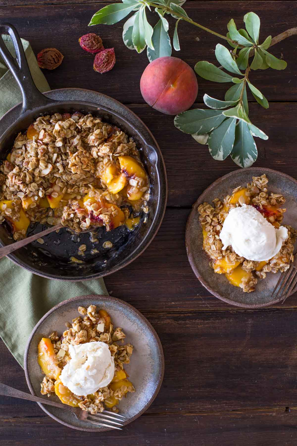 Two plates of Easy Skillet Peach Crisp topped with vanilla ice cream, sitting next to the skillet of Peach Crisp, as well as a whole peach and a few peach pits. 