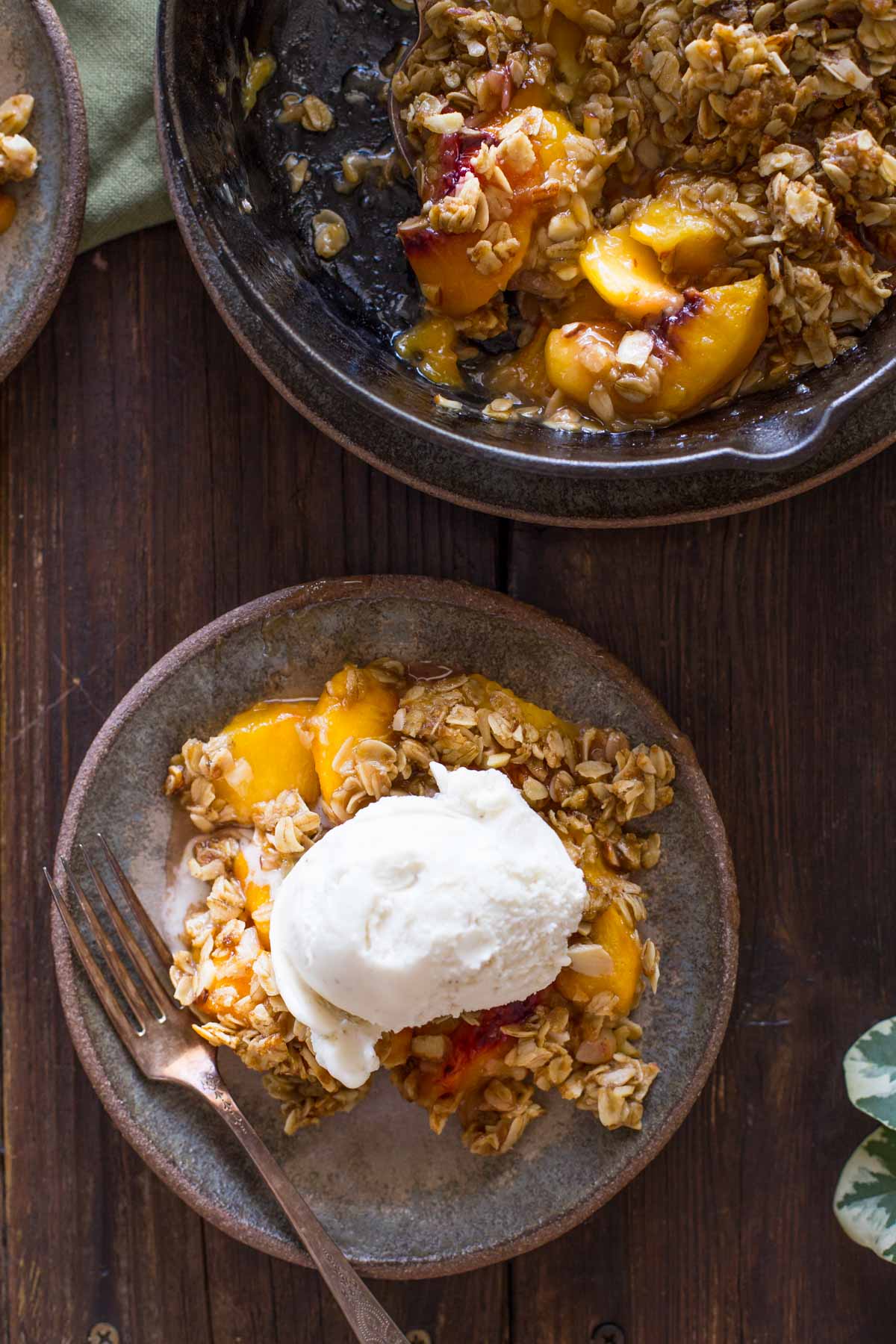 A plate of Easy Skillet Peach Crisp topped with vanilla ice cream, sitting next to the skillet of Peach Crisp.