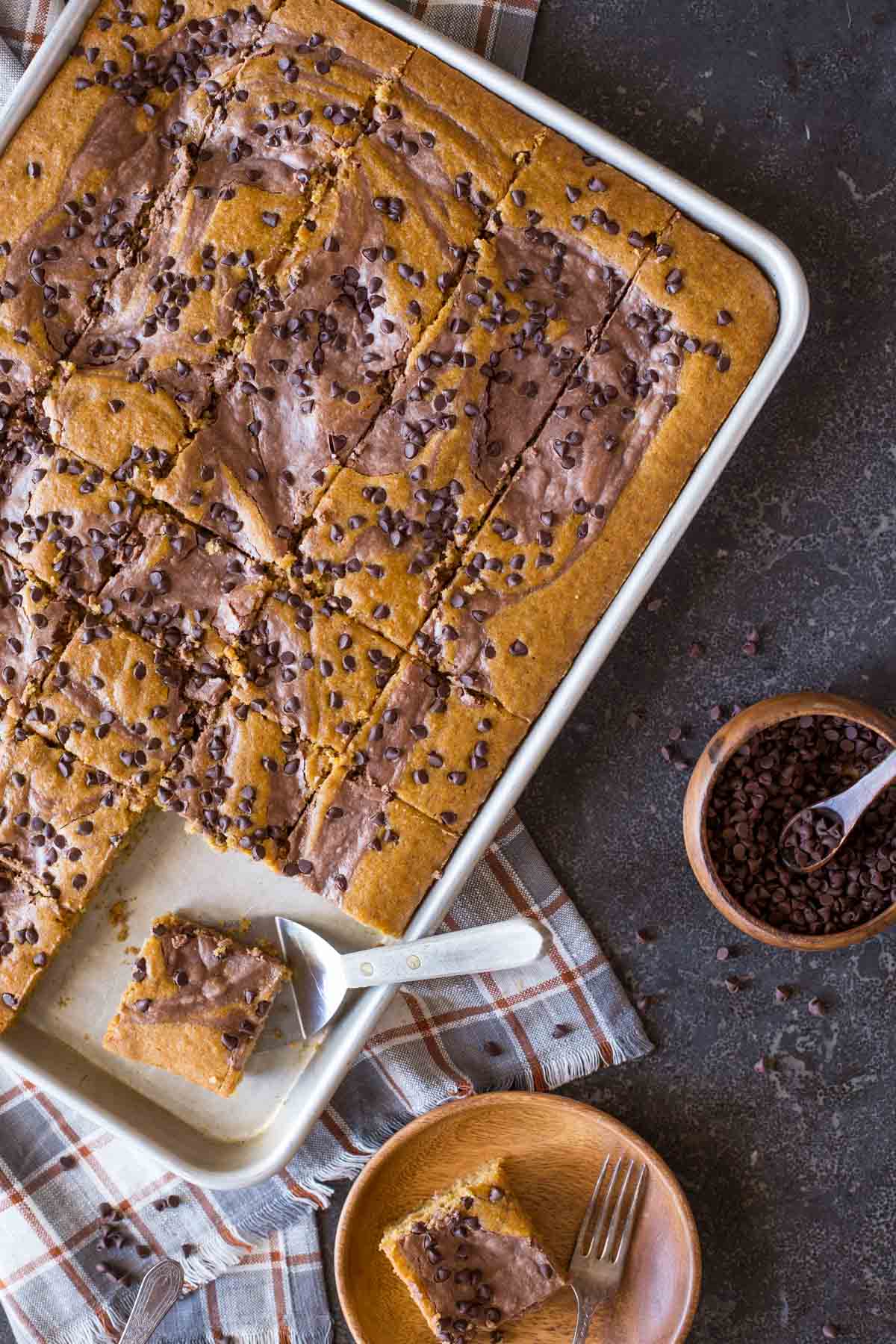 Chocolate-Swirled Pumpkin Bars cut in a baking pan with a serving spatula under one of the bars, and a wood plate sitting next to the pan with a Chocolate-Swirled Pumpkin Bar on it with a fork, and a wood bowl of chocolate chips next to the pan. 
