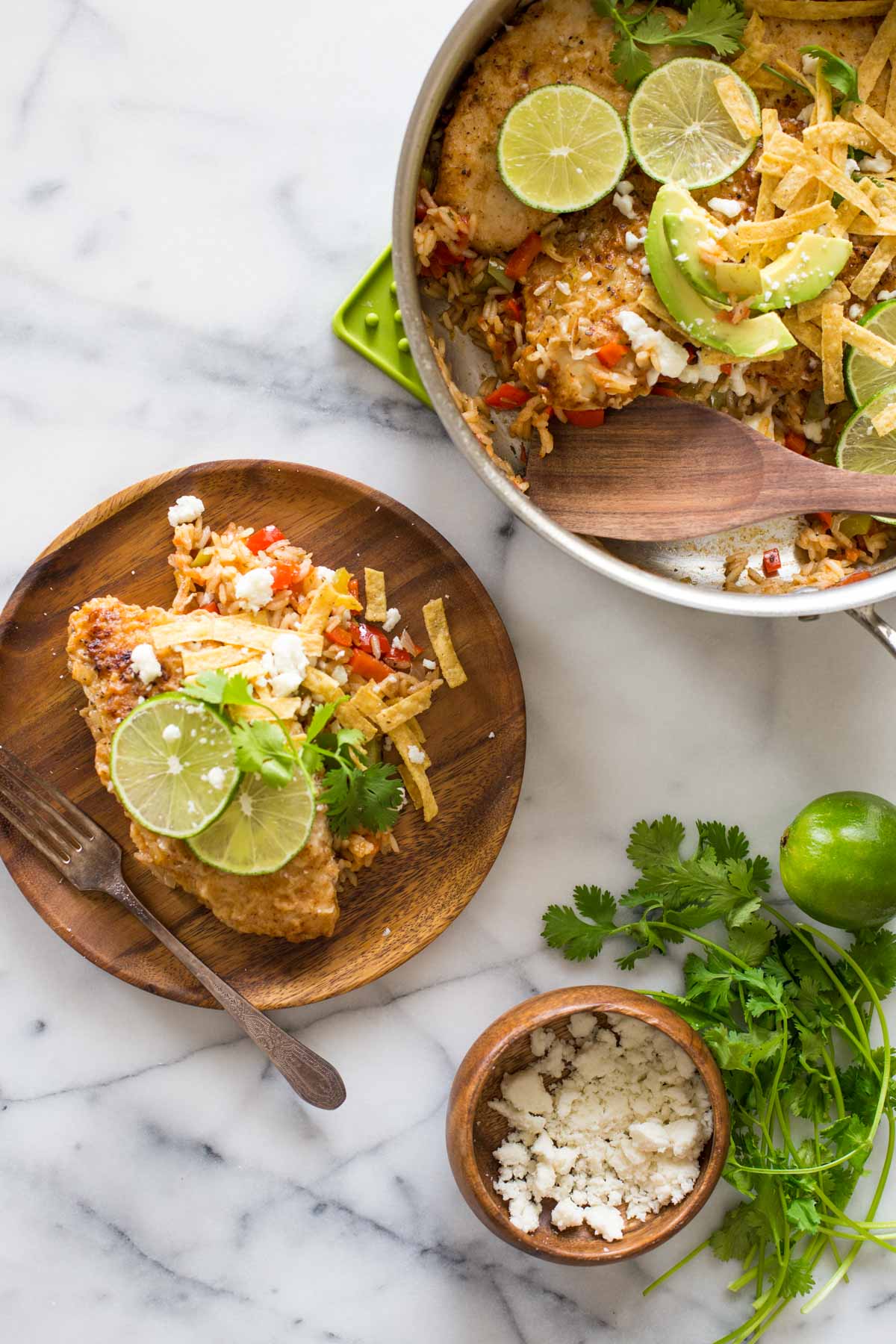 A plate with a serving of One Skillet Fajita Style Chicken and Rice, topped with crumbled queso fresco, fresh cilantro, tortilla strips, and lime slices, sitting next to the skillet of One Skillet Fajita Style Chicken and Rice, and a small wood bowl of crumbled queso fresco, fresh cilantro and a whole lime. 