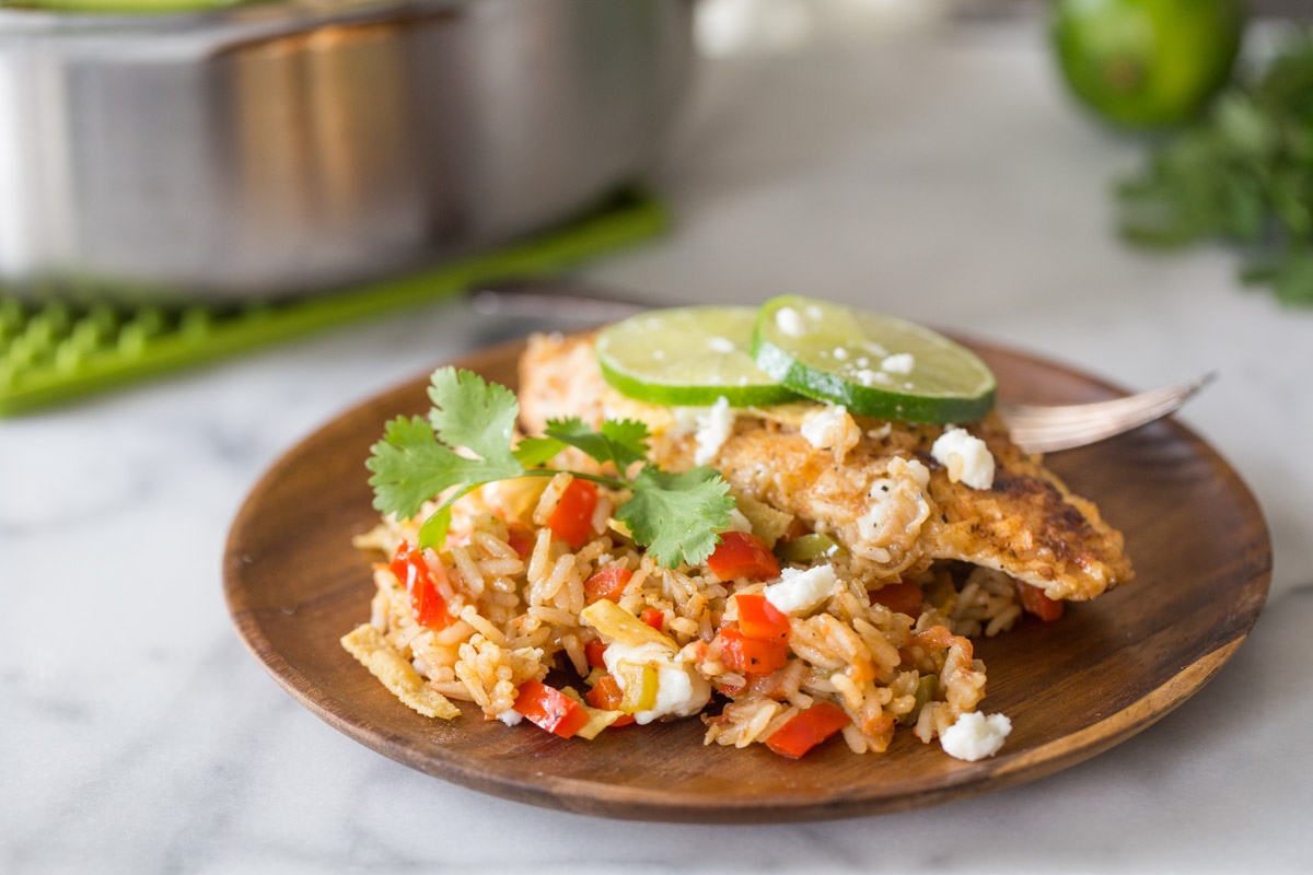 A plate with a serving of One Skillet Fajita Style Chicken and Rice, topped with crumbled queso fresco, fresh cilantro, tortilla strips, and lime slices.
