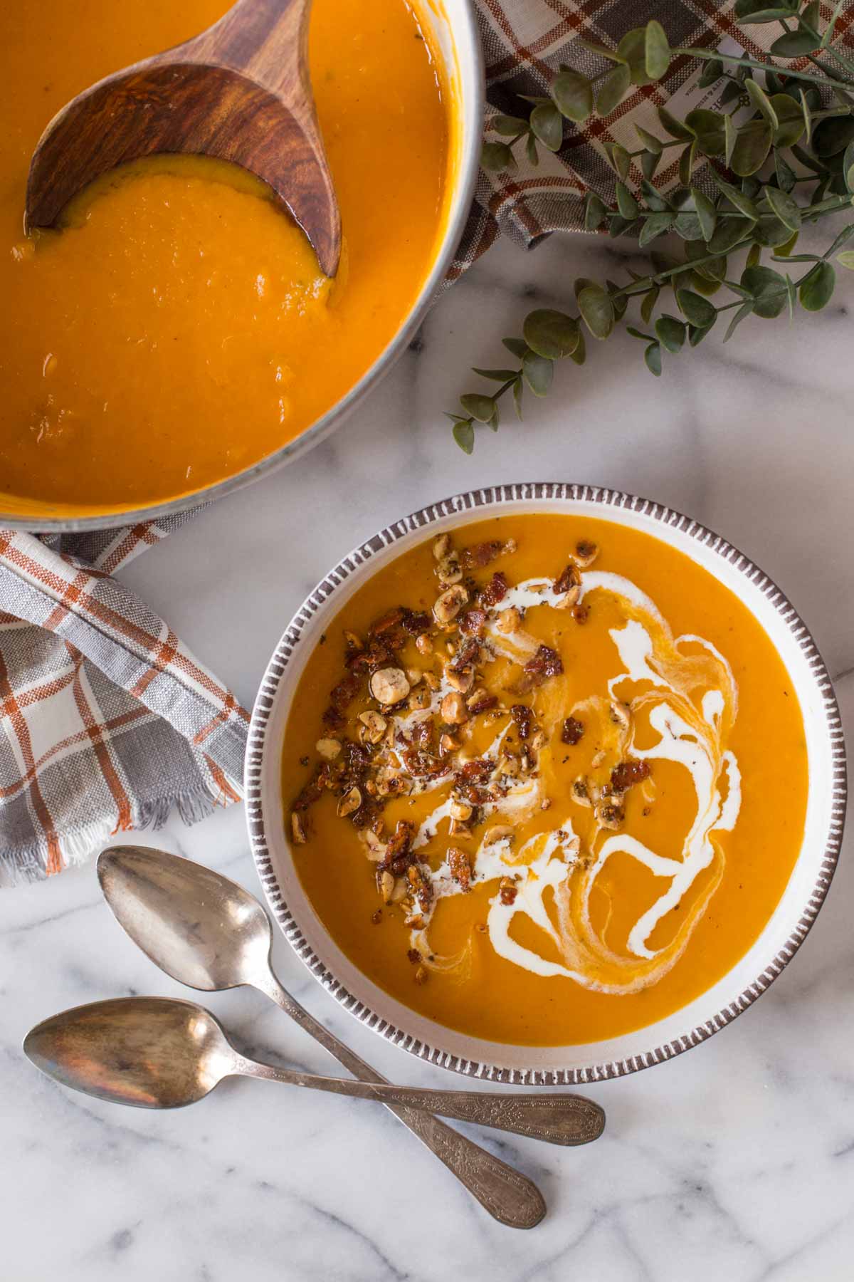 Roasted Butternut Squash Soup in a bowl with a swirl of heavy cream, and topped with a sprinkle of bacon and hazelnuts, sitting next to a pot of Roasted Butternut Squash Soup.