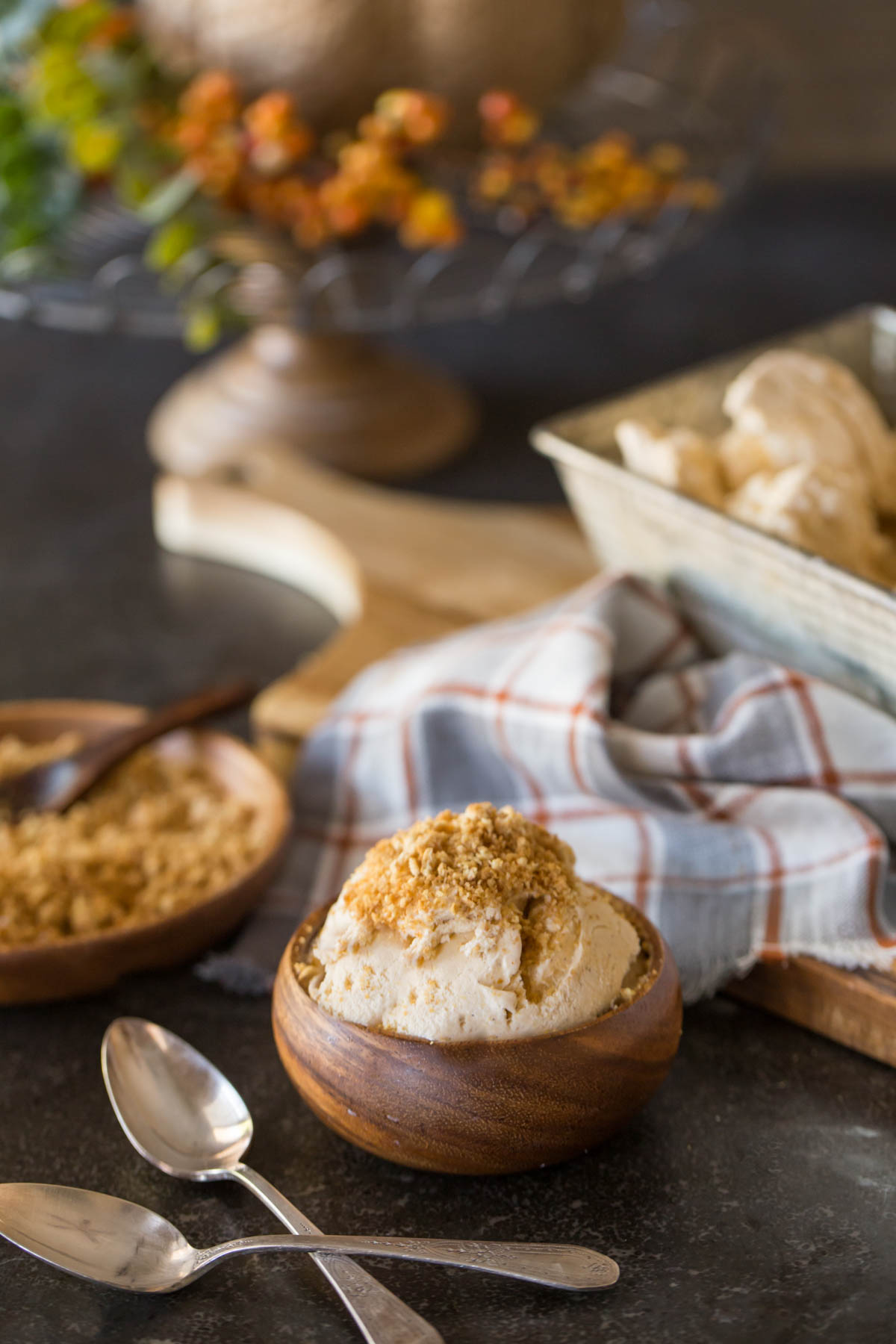 A wood bowl of Homemade Pumpkin Pie Ice Cream topped with crushed graham cracker crumbs, with two spoons sitting next to it, and a wood plate of crushed graham cracker crumbs in the background, along with the loaf pan container of Homemade Pumpkin Pie Ice Cream. 