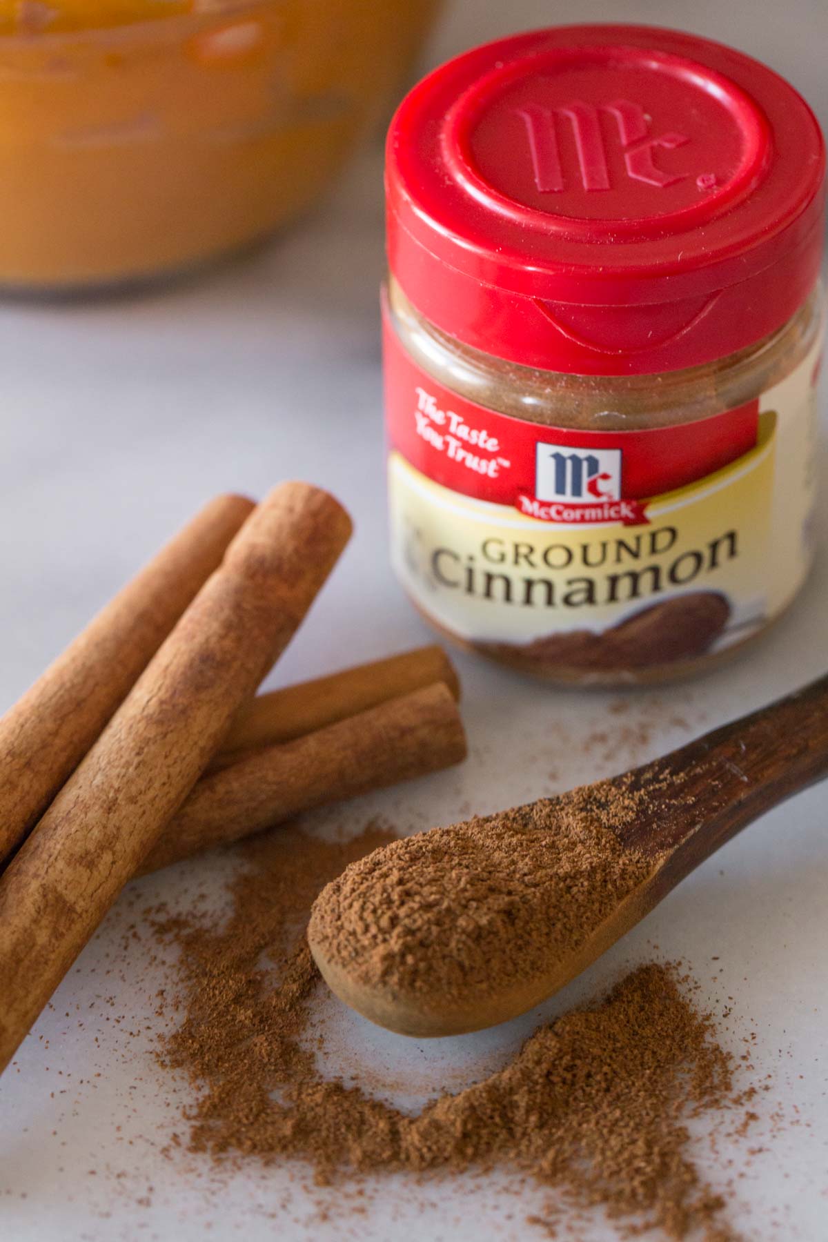 A McCormick spice container of Ground Cinnamon, with a few cinnamon sticks and a small wood spoon of ground cinnamon sitting in front of it.  
