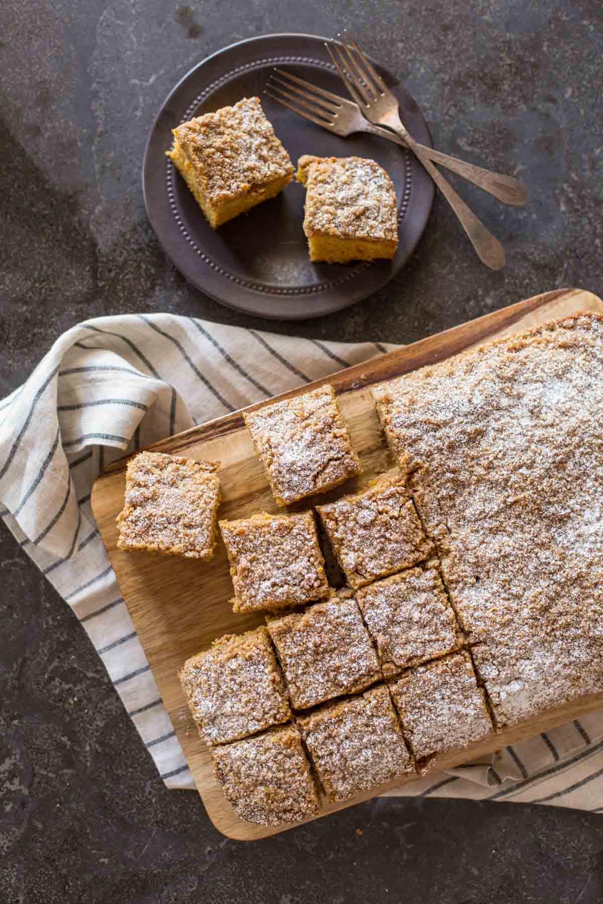 Pumpkin Spice Coffee Cake on a cutting board, with half of the cake cut into square servings, and a plate with two pieces of the cake and two forks sitting next to the cutting board. 