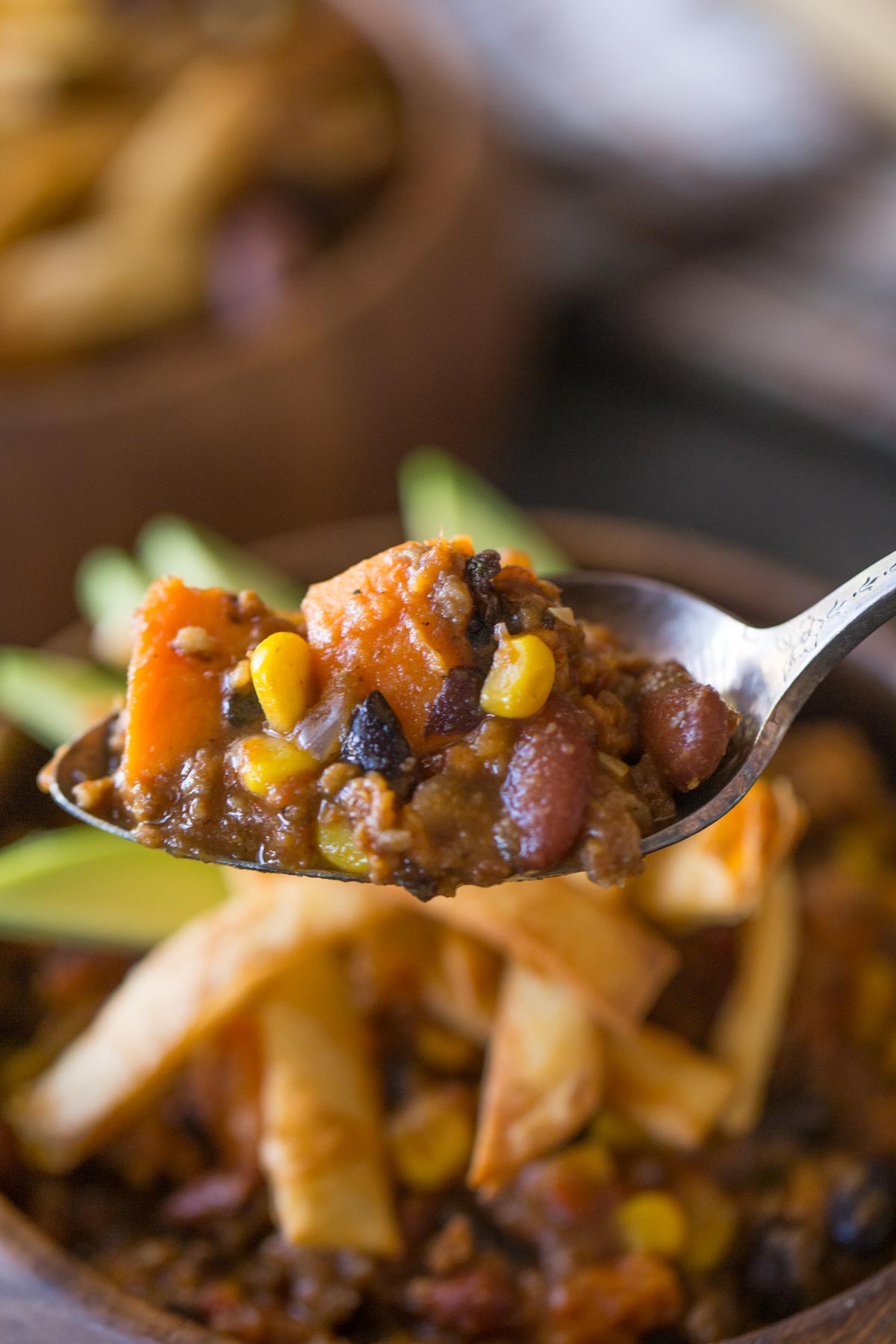 A spoon full of Sweet Potato Chili, with a wood bowl of Sweet Potato Chili, topped with Fried Tortilla Strips and sliced avocado in the background. 