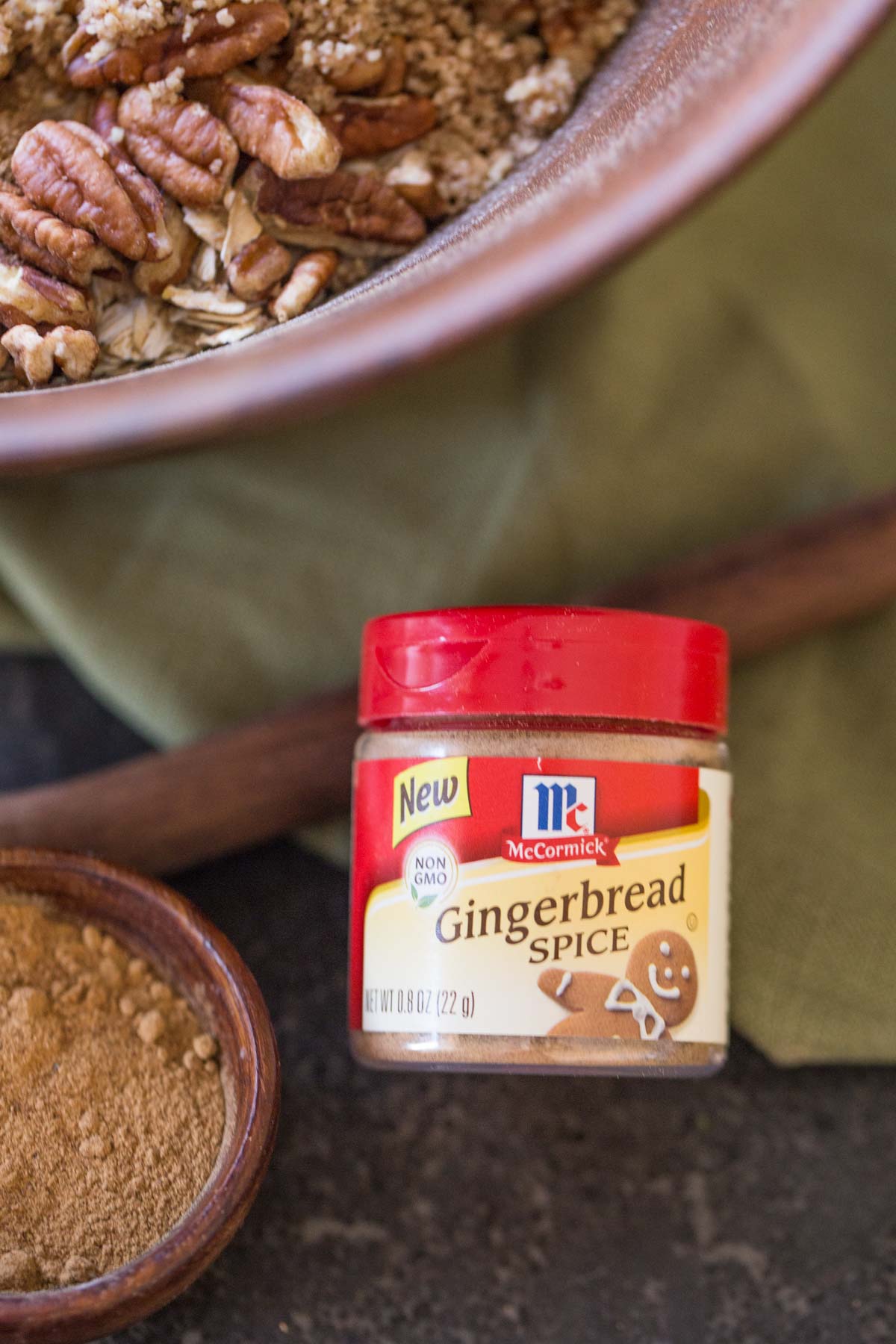 A container of McCormick Gingerbread Spice mix, sitting next to a small wood bowl of the spice mix and a large bowl of the Gingerbread Spice Granola ingredients. 
