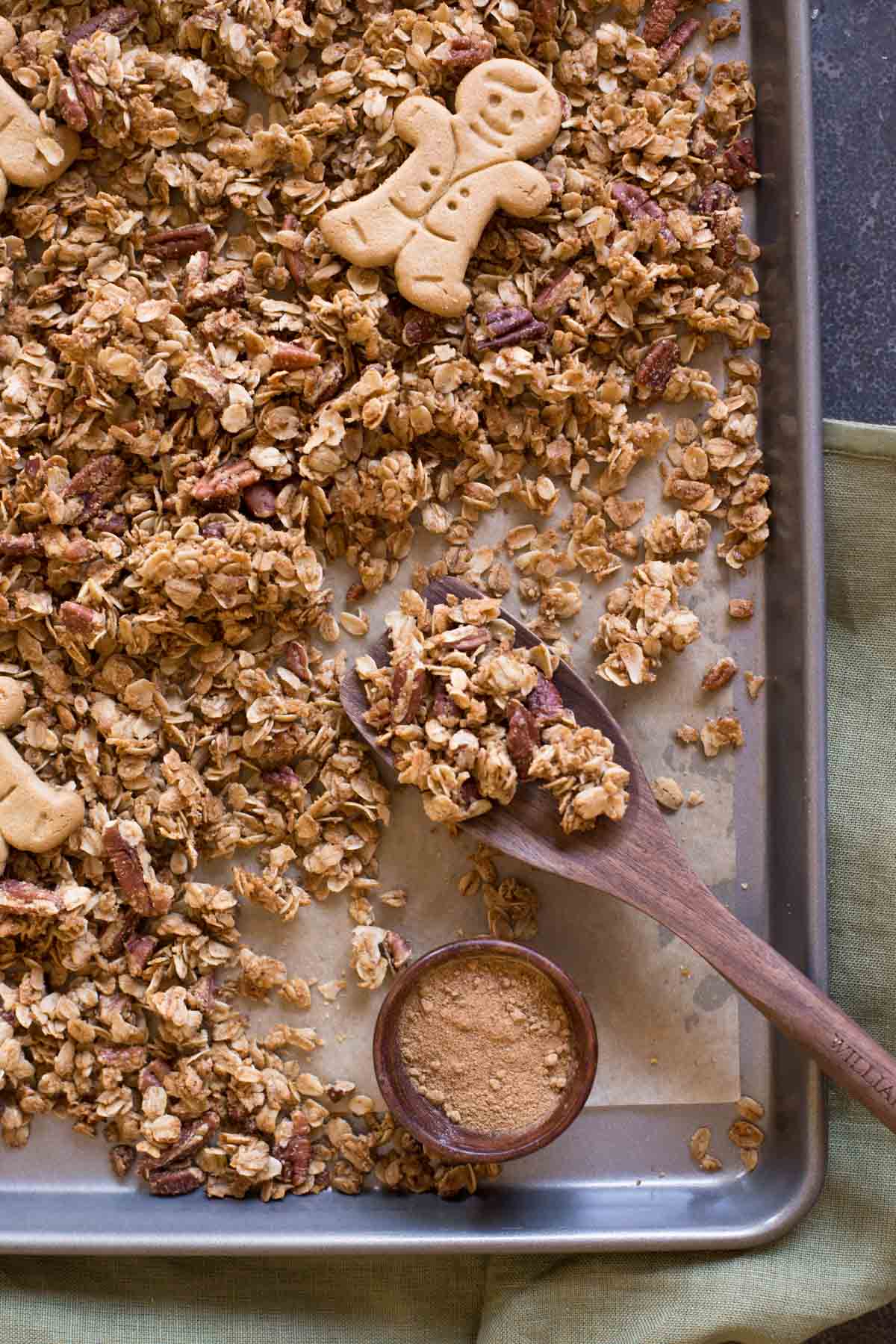 Gingerbread Spice Granola on a parchment paper lined baking sheet with whole gingerbread man cookies on top of the granola and a small wood bowl of the spices sitting on the baking sheet, along with a wooden spoon. 