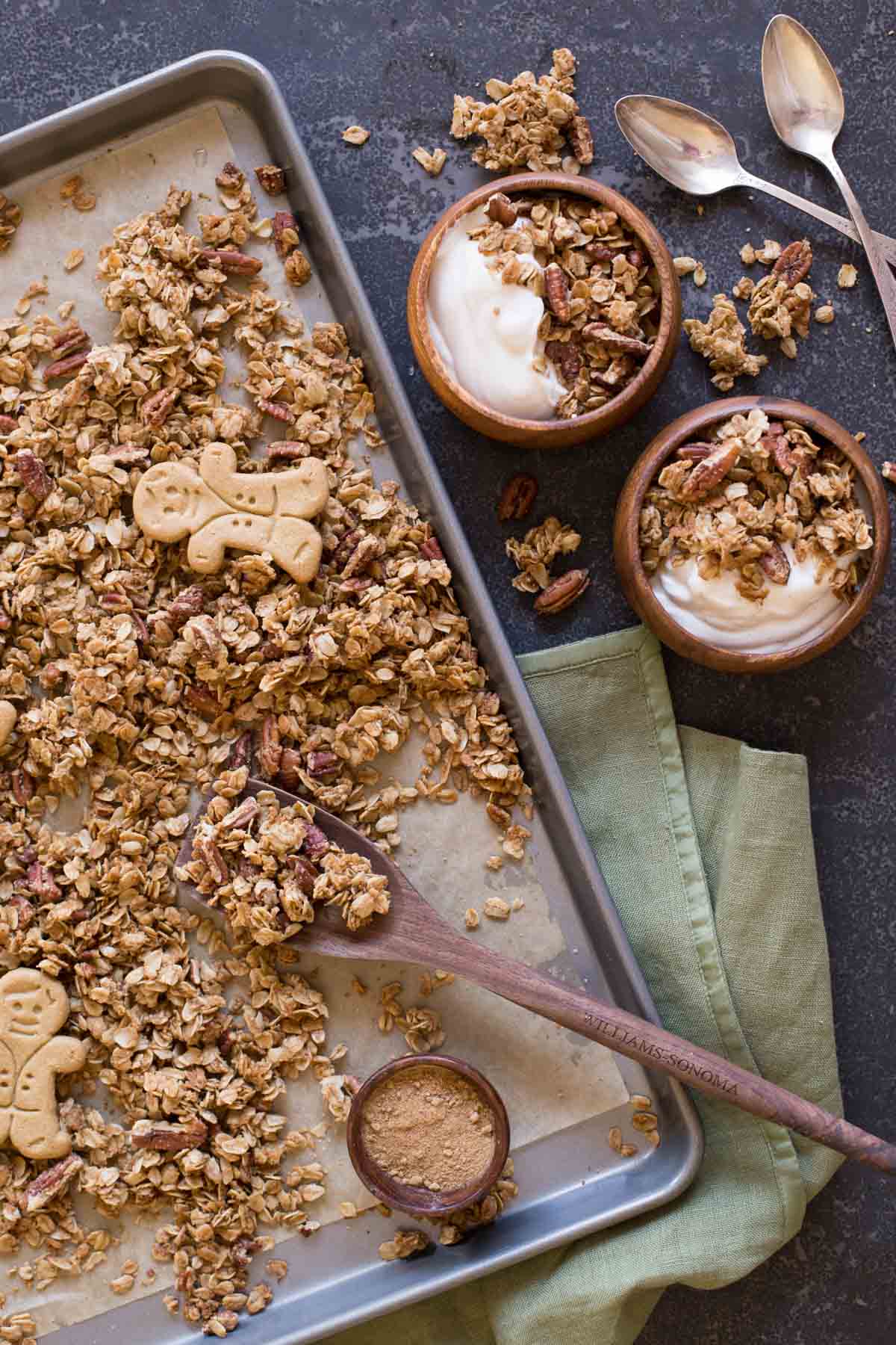 Gingerbread Spice Granola on a parchment paper lined baking sheet with two whole gingerbread man cookies on top of the granola and a small wood bowl of the spices sitting on the baking sheet, along with a wooden spoon.  Next to the baking sheet are two bowls of yogurt and the Gingerbread Spice Granola and two spoons.  