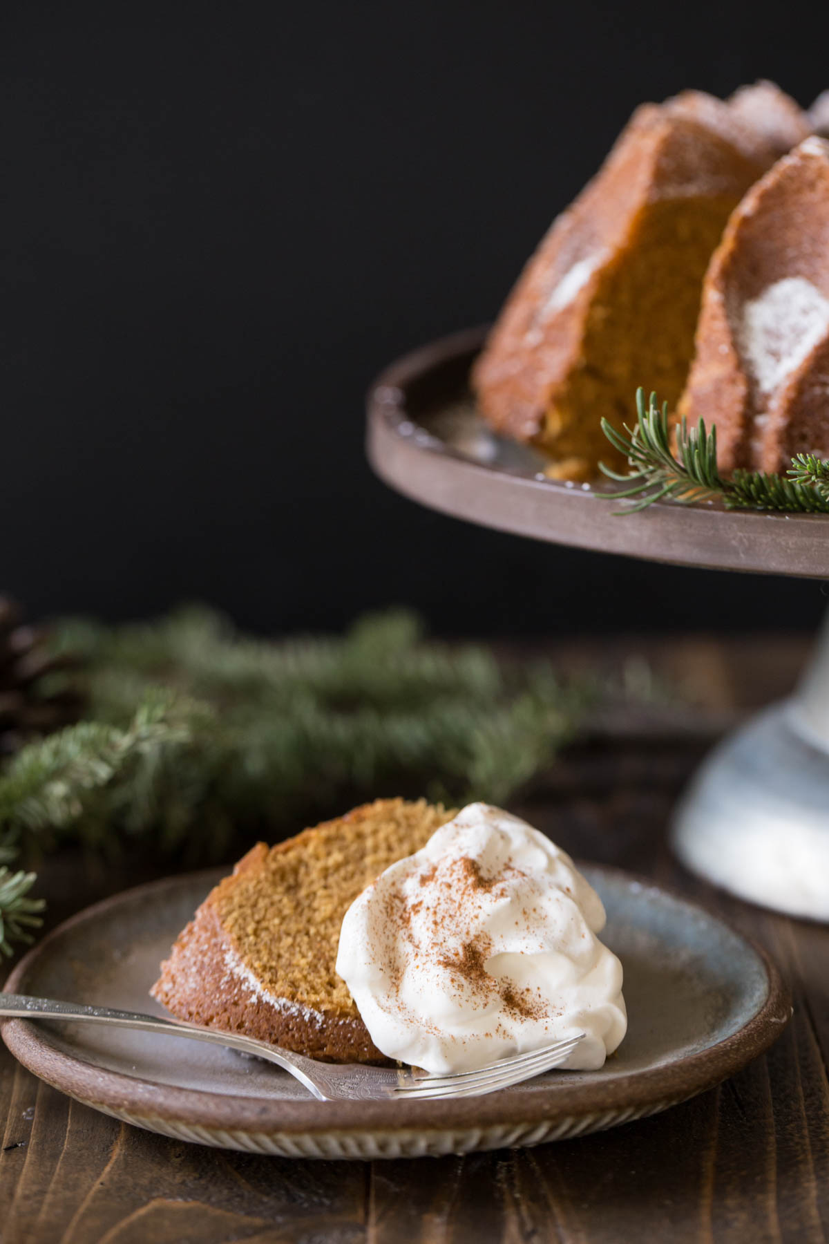 A slice of Gingerbread Bundt Cake on a plate, topped with whipped cream, sitting next to the rest of the Gingerbread Bundt Cake on a cake stand.