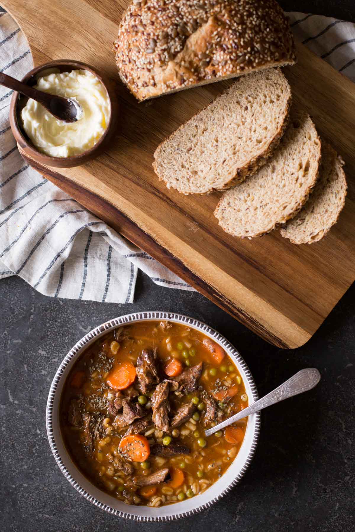 Beef and Barley Vegetable Soup in a bowl with a spoon, sitting next to a cutting board with bread and a small bowl of soft butter. 