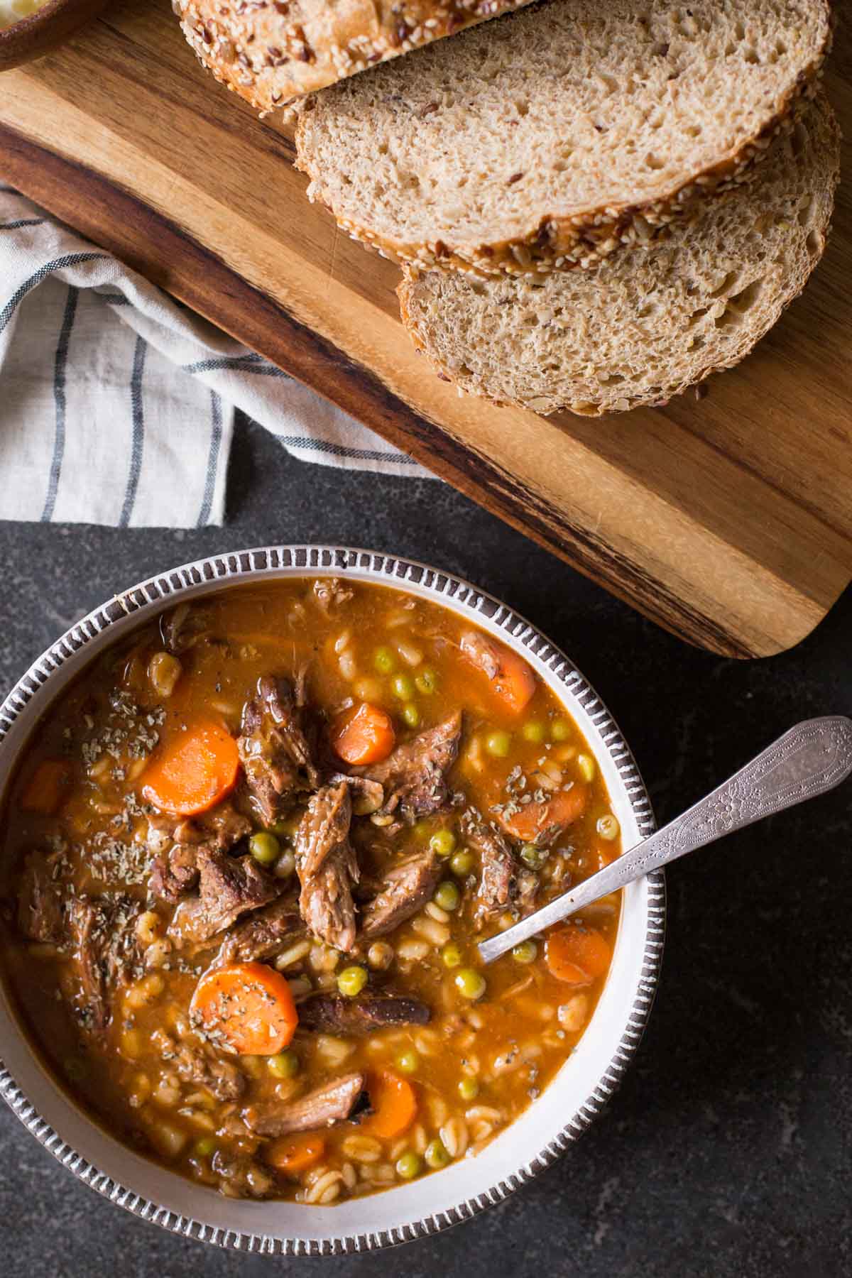 Beef and Barley Vegetable Soup in a bowl with a spoon, sitting next to a cutting board with bread on it. 