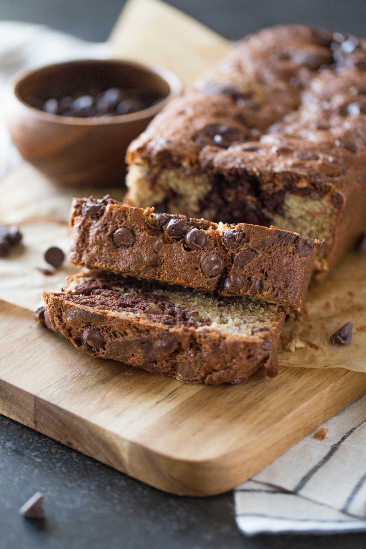 A loaf of Marbled Chocolate Banana Bread on a cutting board with a small wood bowl of chocolate chips. 