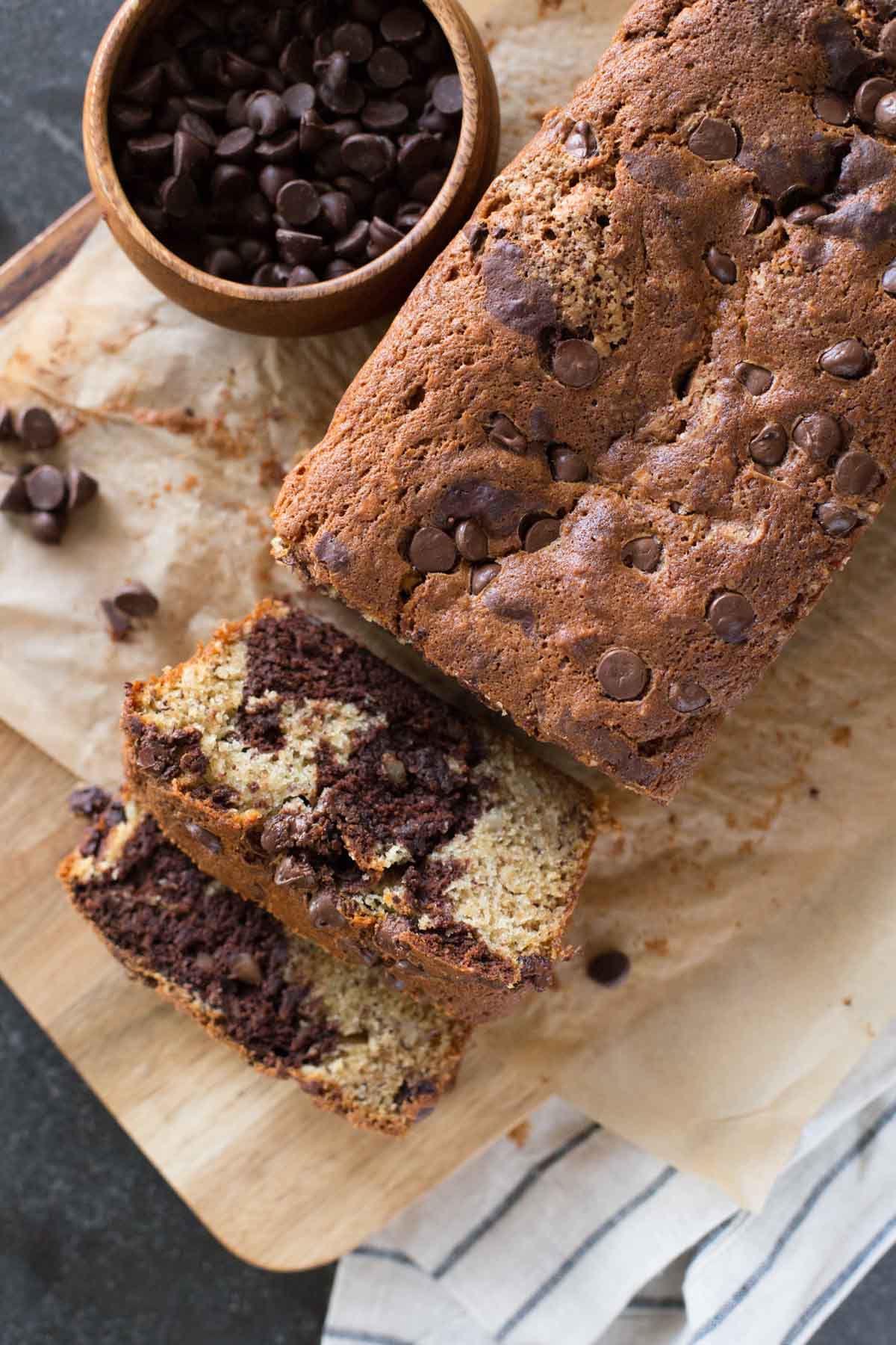 A loaf of Marbled Chocolate Banana Bread on a cutting board with a small wood bowl of chocolate chips. 