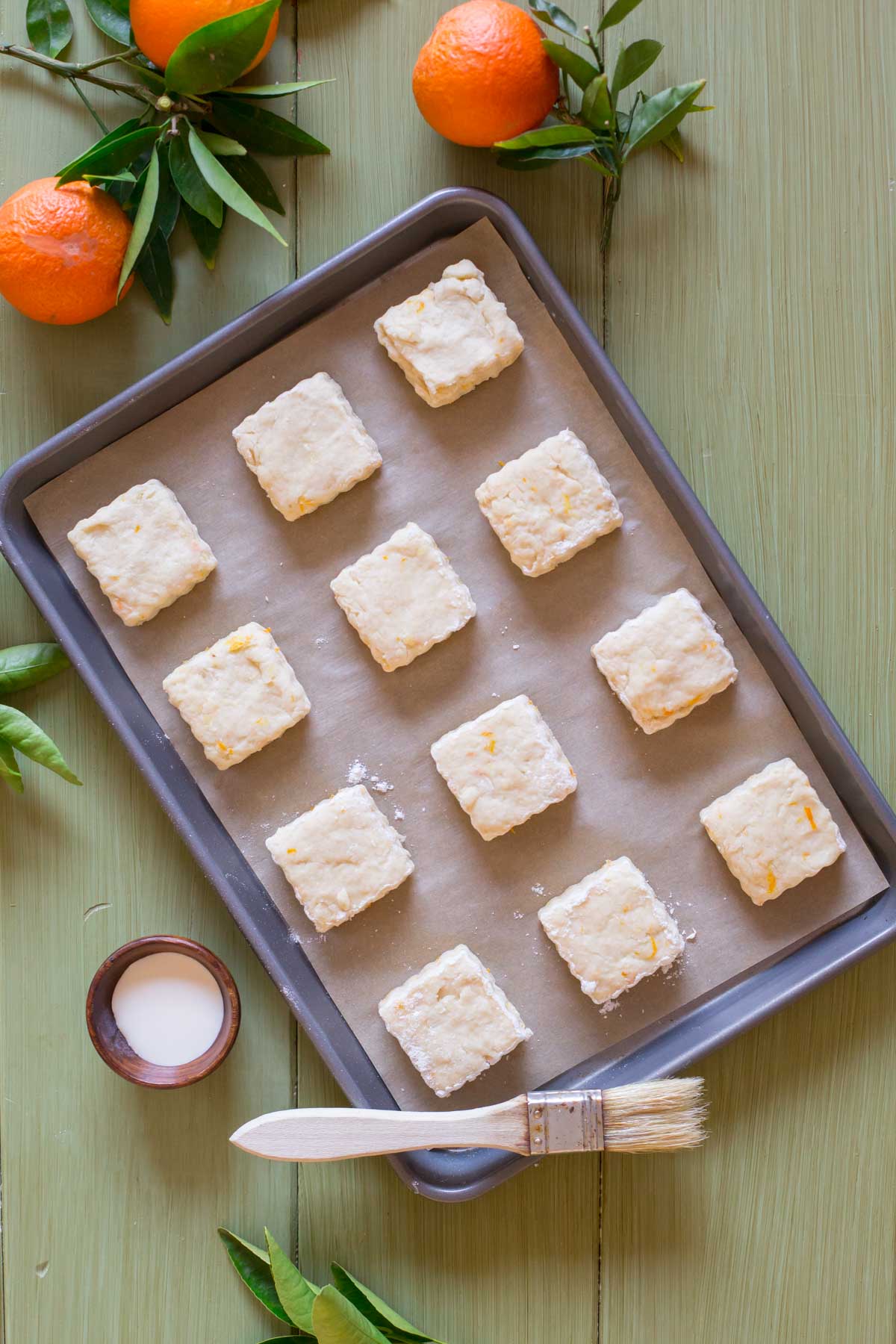 Mini Orange Cream Scones on a parchment paper lined baking sheet, with a small bowl of heavy cream next to it, along with a pastry brush. 