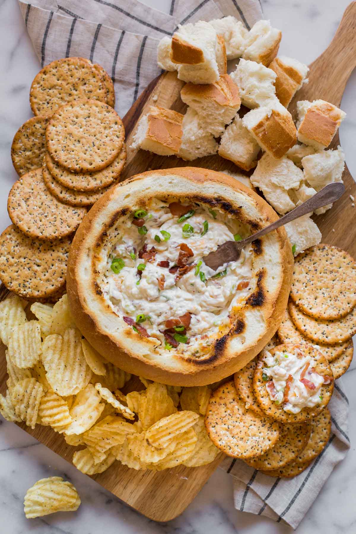 Three Cheese Bacon Dip in a bread bowl sitting on a wood board, surrounded by bread chunks, chips and crackers. 