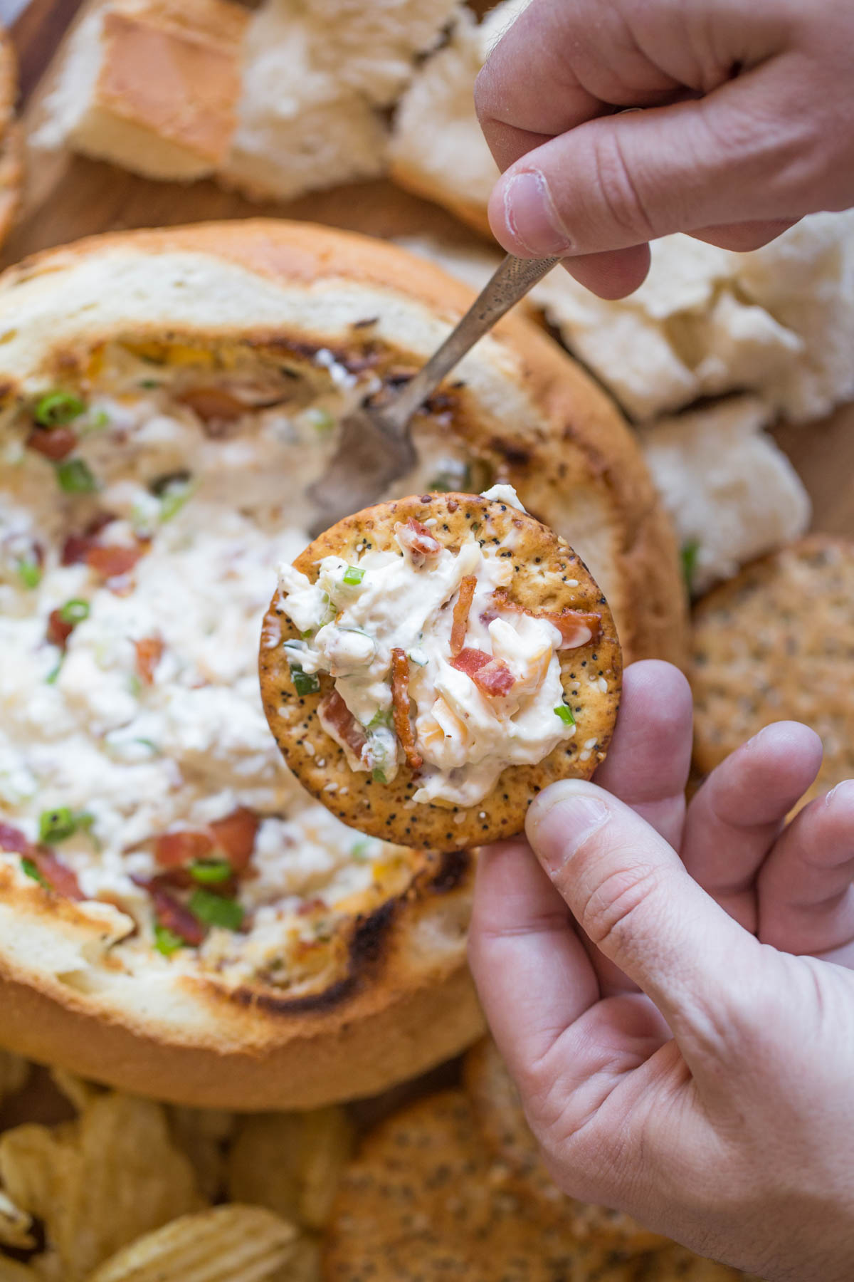 Three Cheese Bacon Dip on a cracker, with the dip in a bread bowl surrounded by bread chunks, chips and crackers in the background. 